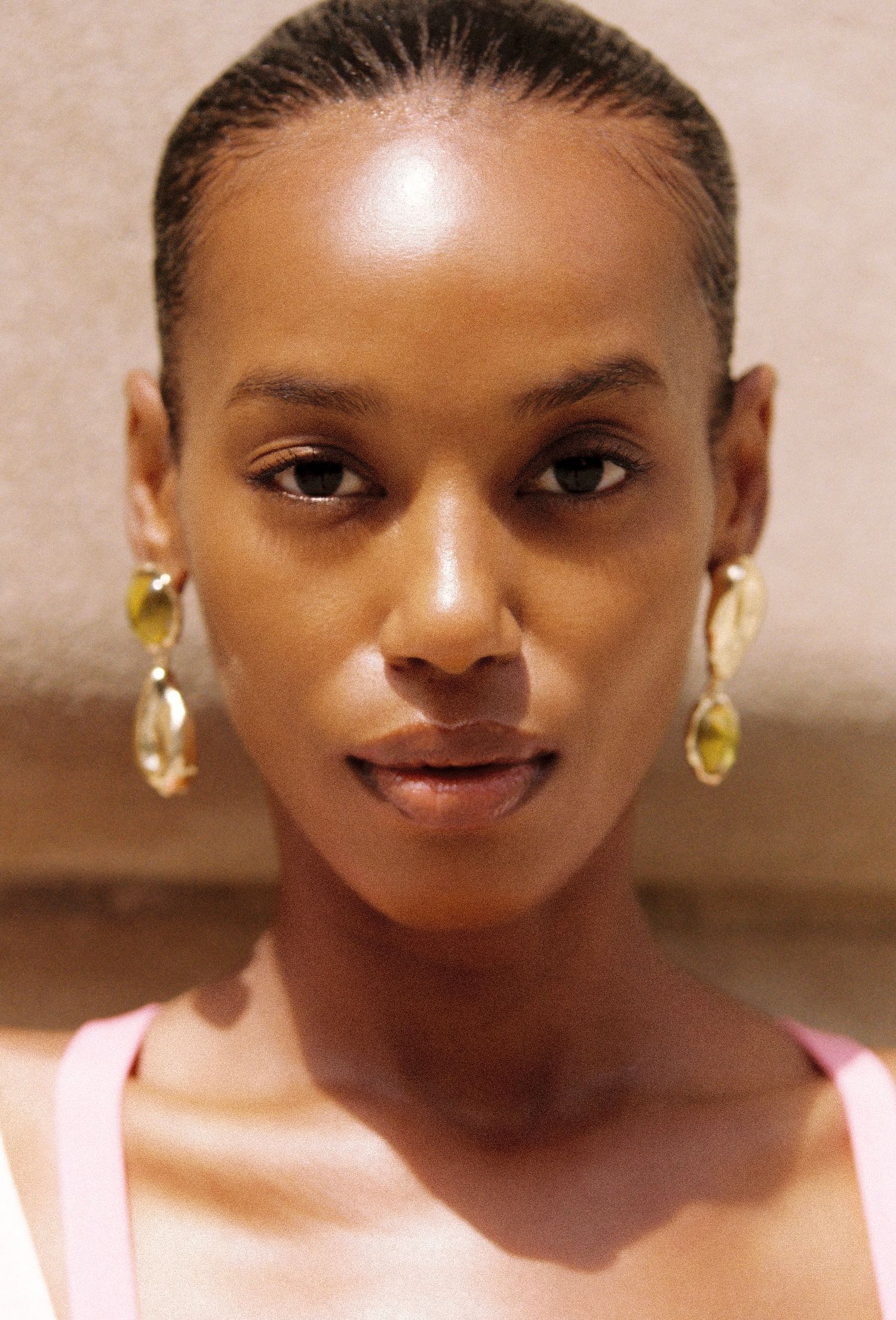 Close-up portrait of a woman with slicked-back hair, wearing gold and olive-green drop earrings and a pink top against a warm beige wall.