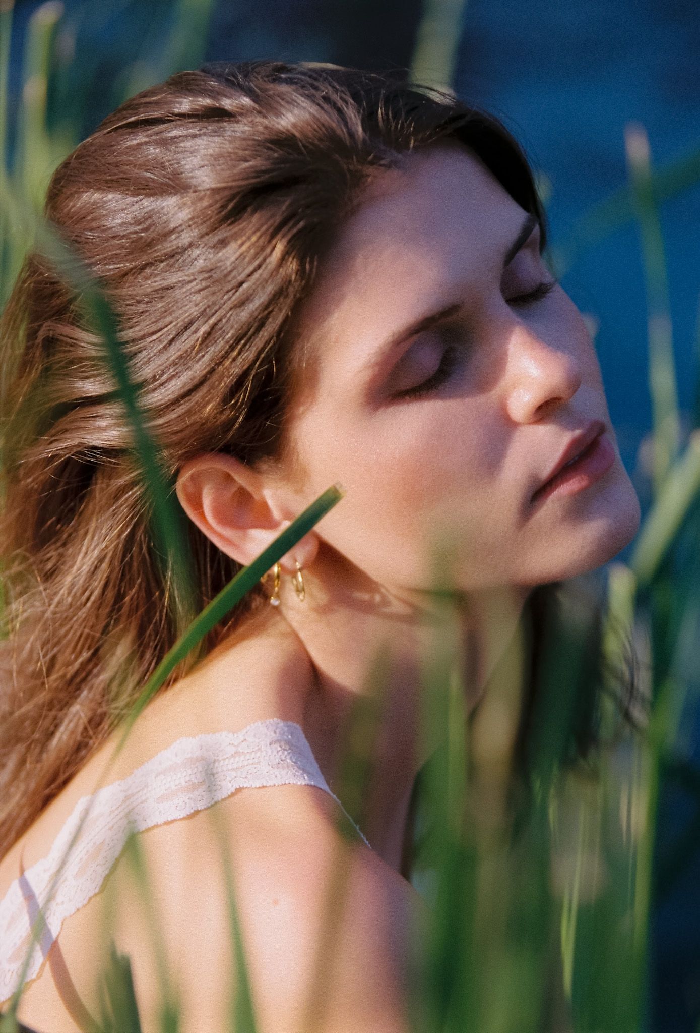 Woman with long brown hair and gold earrings closes her eyes in warm sunlight among tall green grass blades by the water.