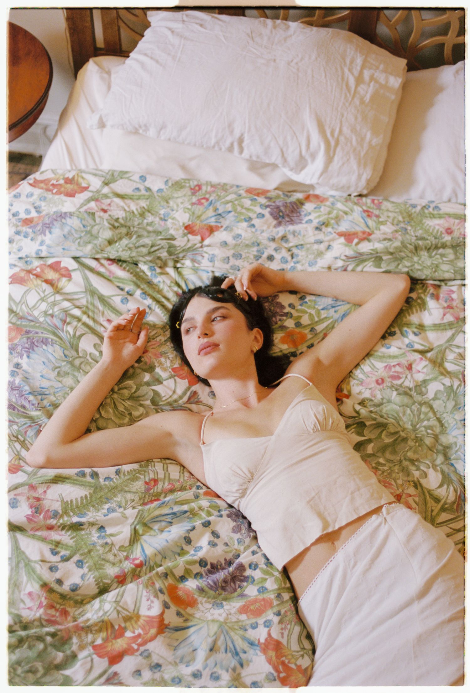 Woman in a cream camisole top and skirt lying on a bed with a floral botanical print comforter and white pillows.