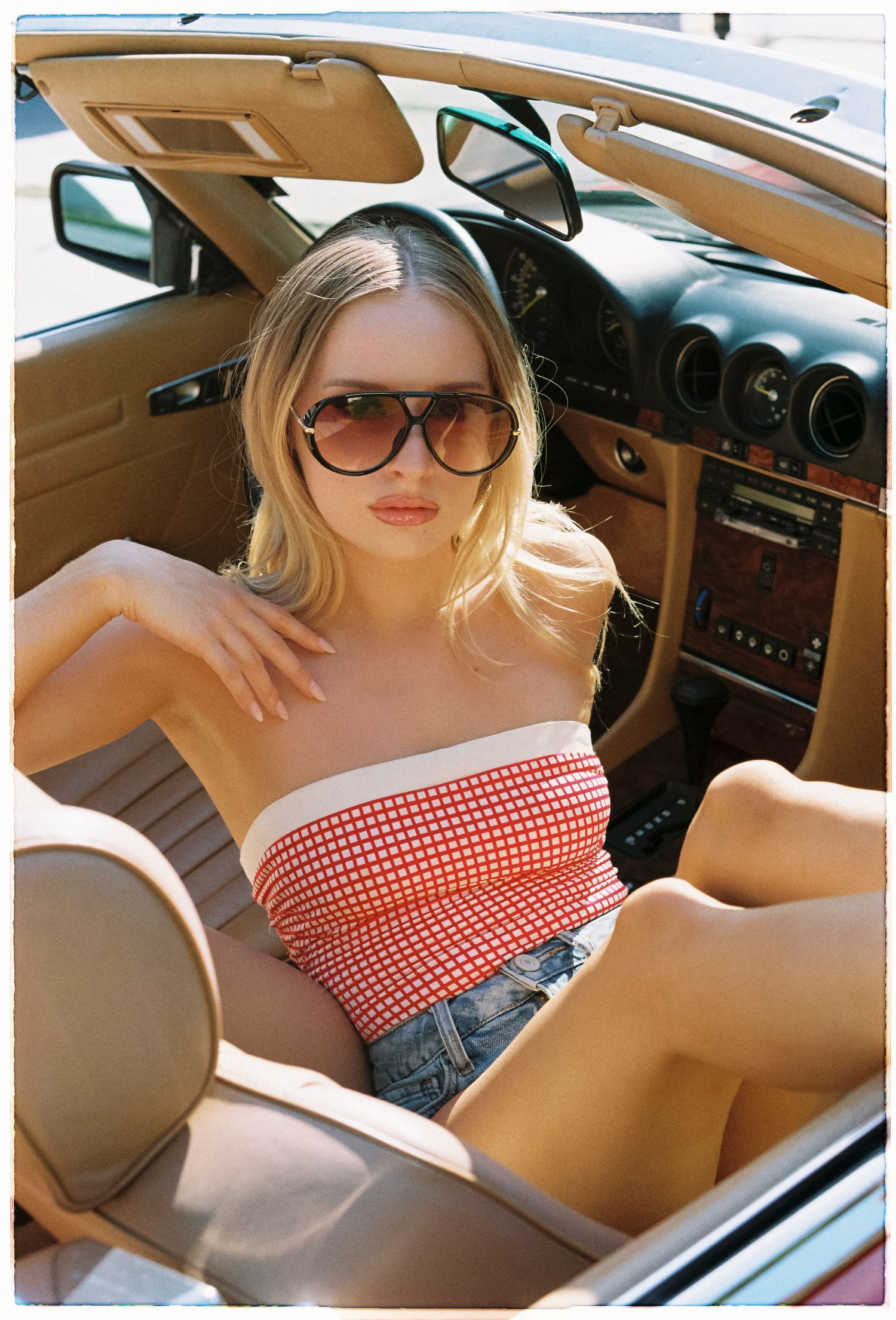 Blonde woman in red gingham tube top and denim shorts wearing oversized sunglasses sitting in a vintage convertible car.