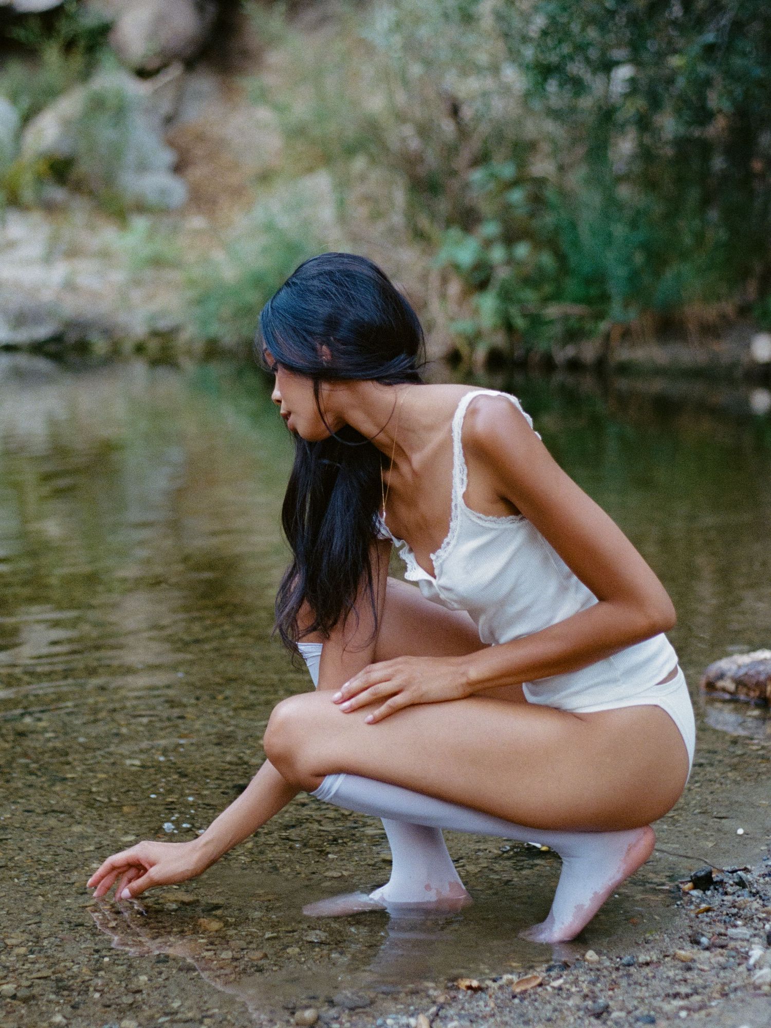 Person in white clothing crouching near water's edge in natural setting.