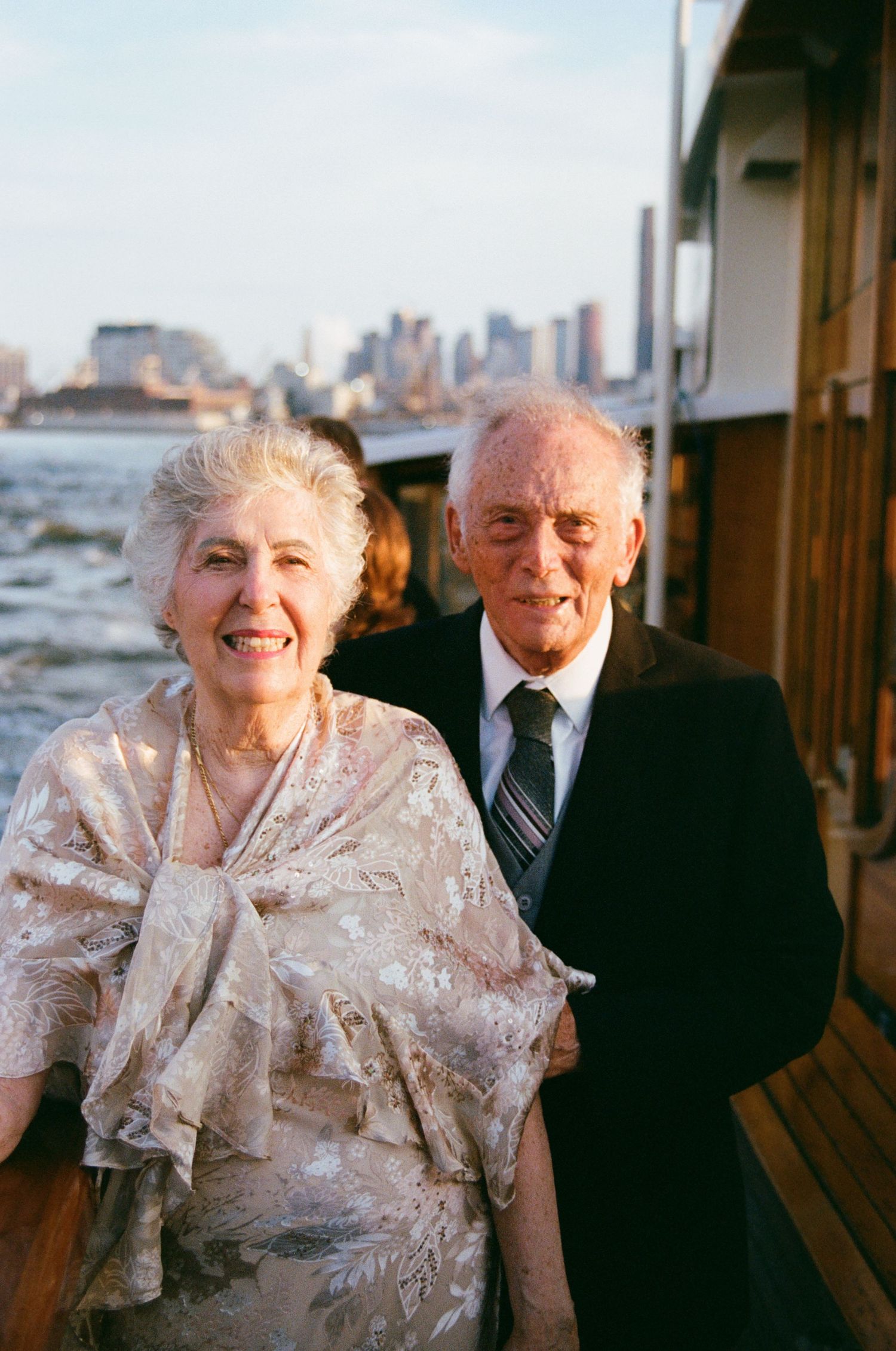 An elderly couple poses together on a boat deck with a city skyline in the background.