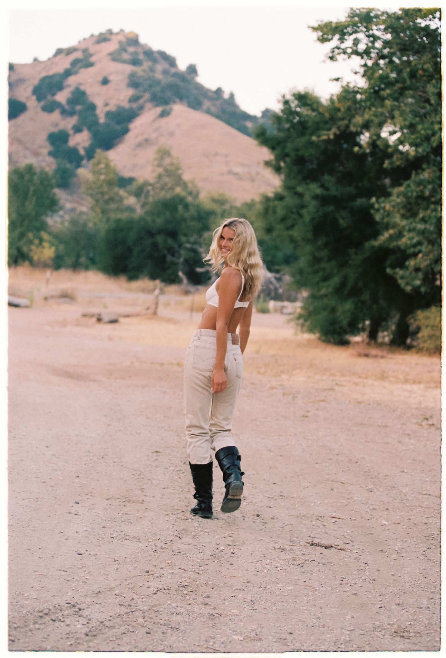 Woman with long blonde hair in a white top, beige pants and black boots looks back while walking on a dirt road near green trees and hills.