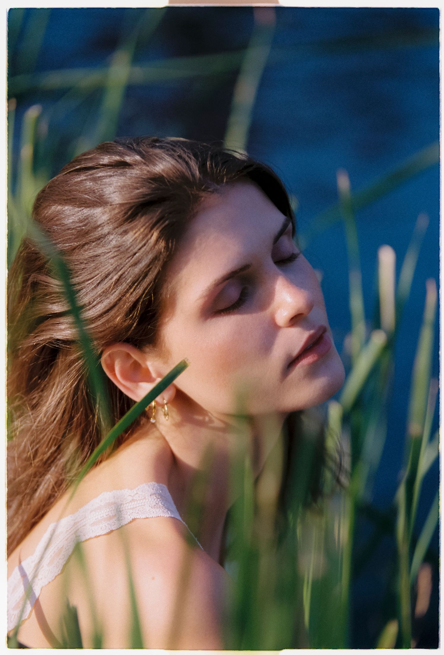 Young woman with brown hair and gold ear cuff poses eyes closed among green reeds near water in soft sunlight.