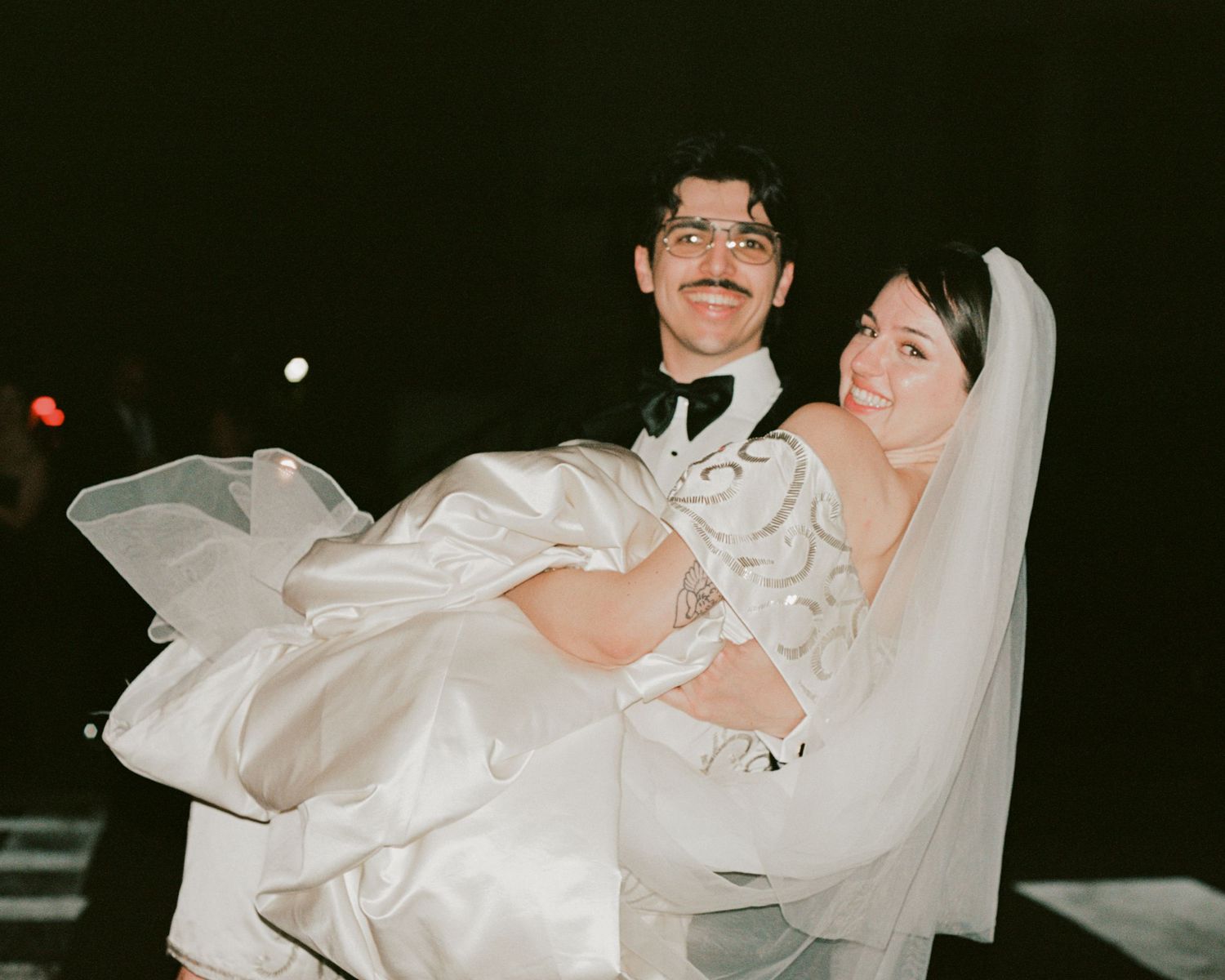 A groom carries his bride in her flowing white dress during nighttime wedding celebrations.