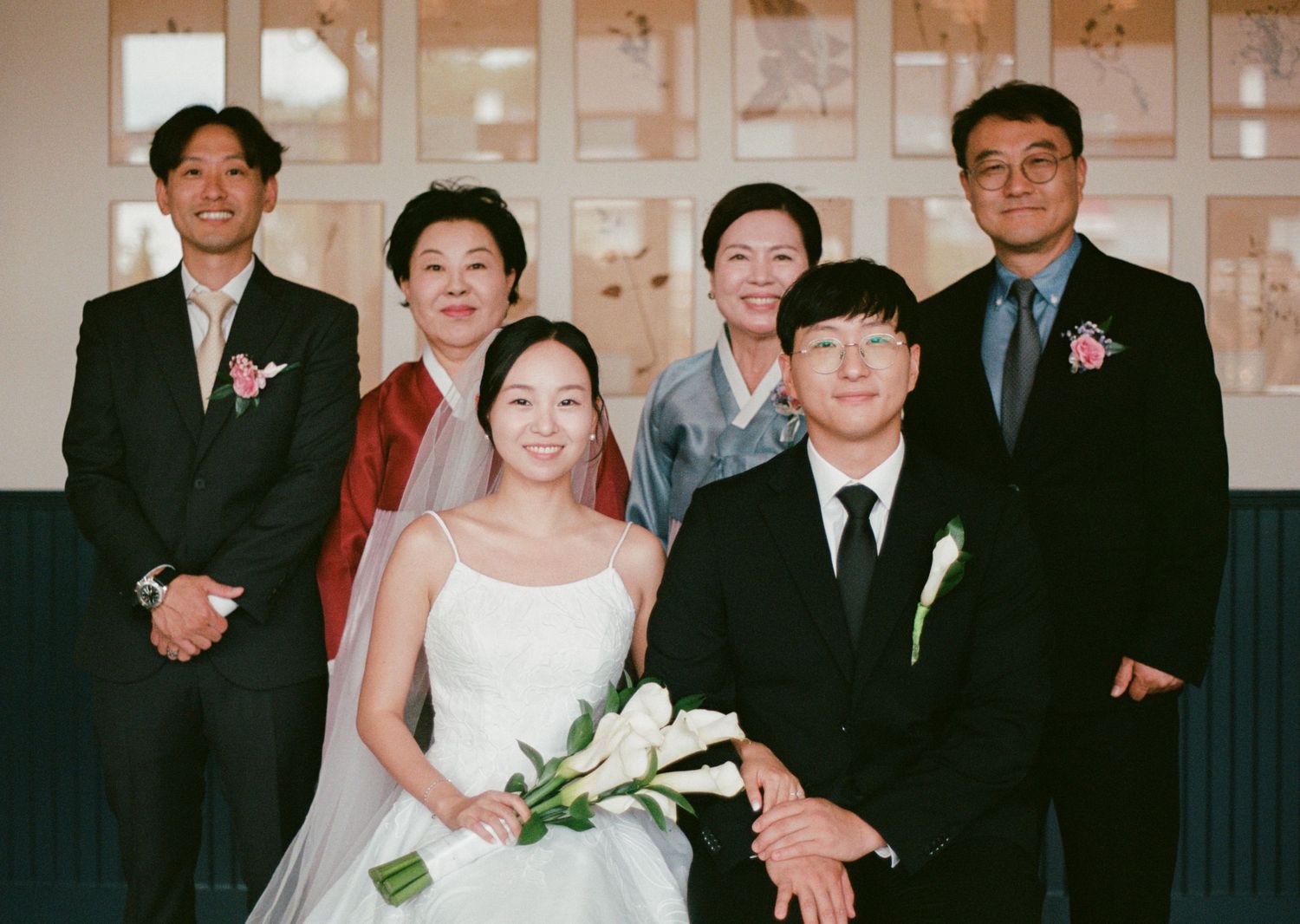 Wedding party poses together in formal attire with bride in white dress holding calla lilies.