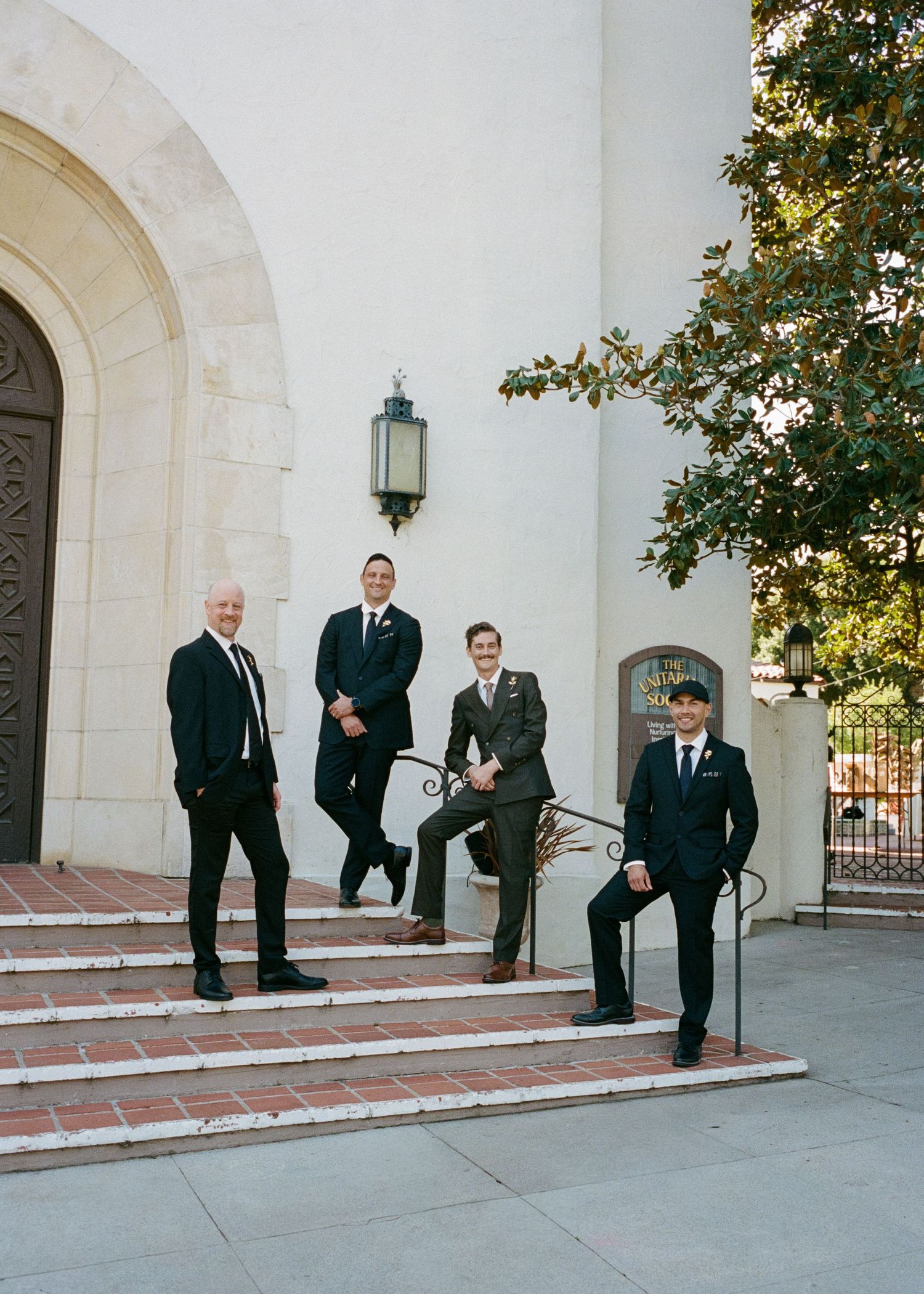 Four groomsmen in dark suits pose casually on brick steps outside a church entrance.