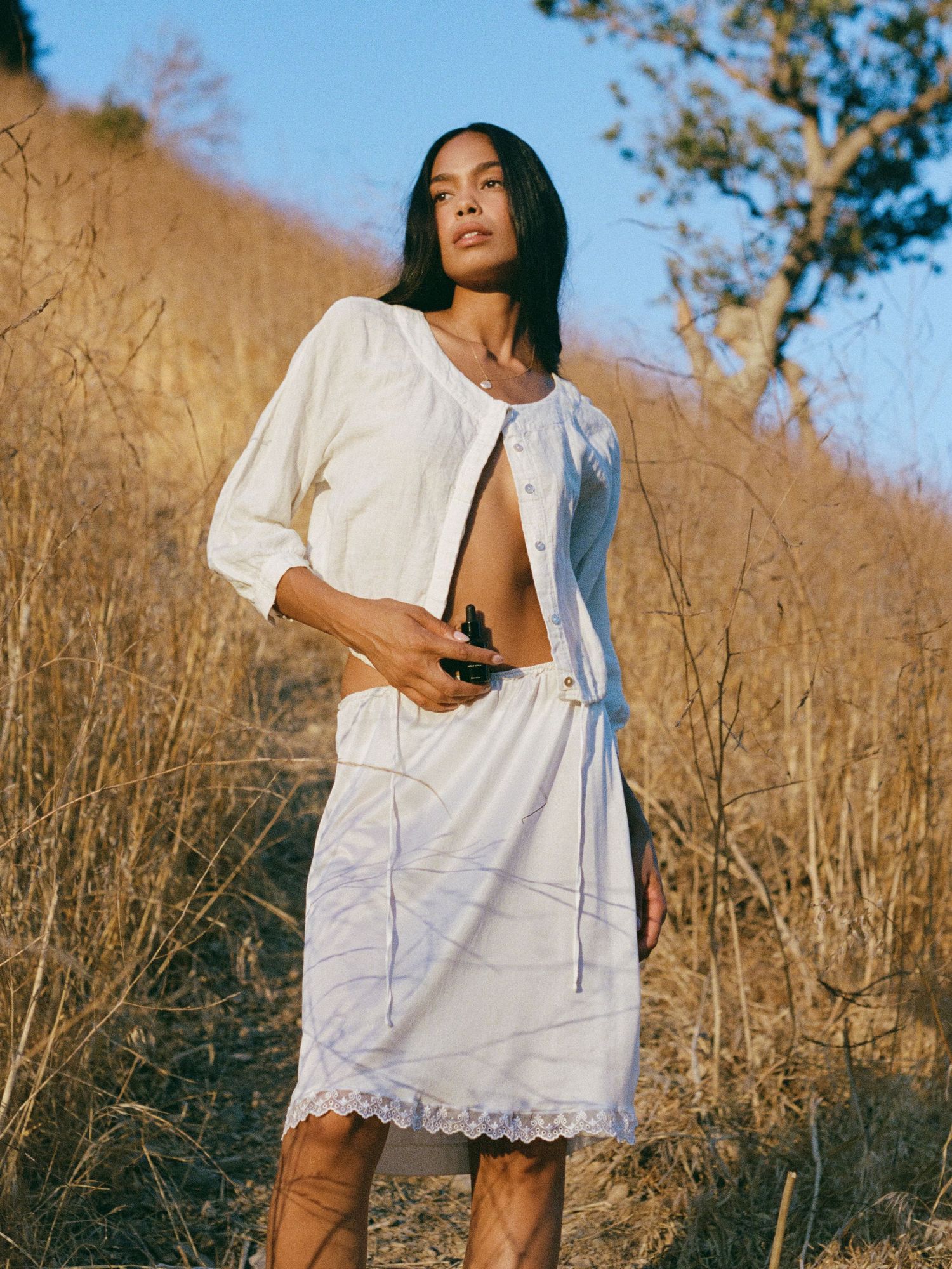 Model wearing white lace dress poses in golden field during sunset.