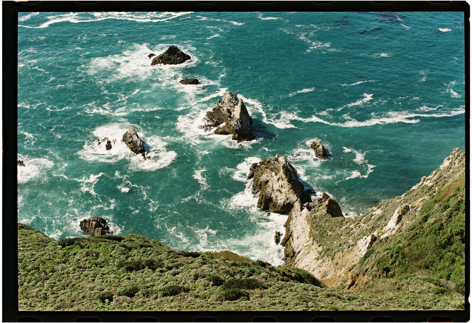 Aerial view of rugged rocky coastline with turquoise ocean waves crashing against jagged sea stacks and green cliffs.