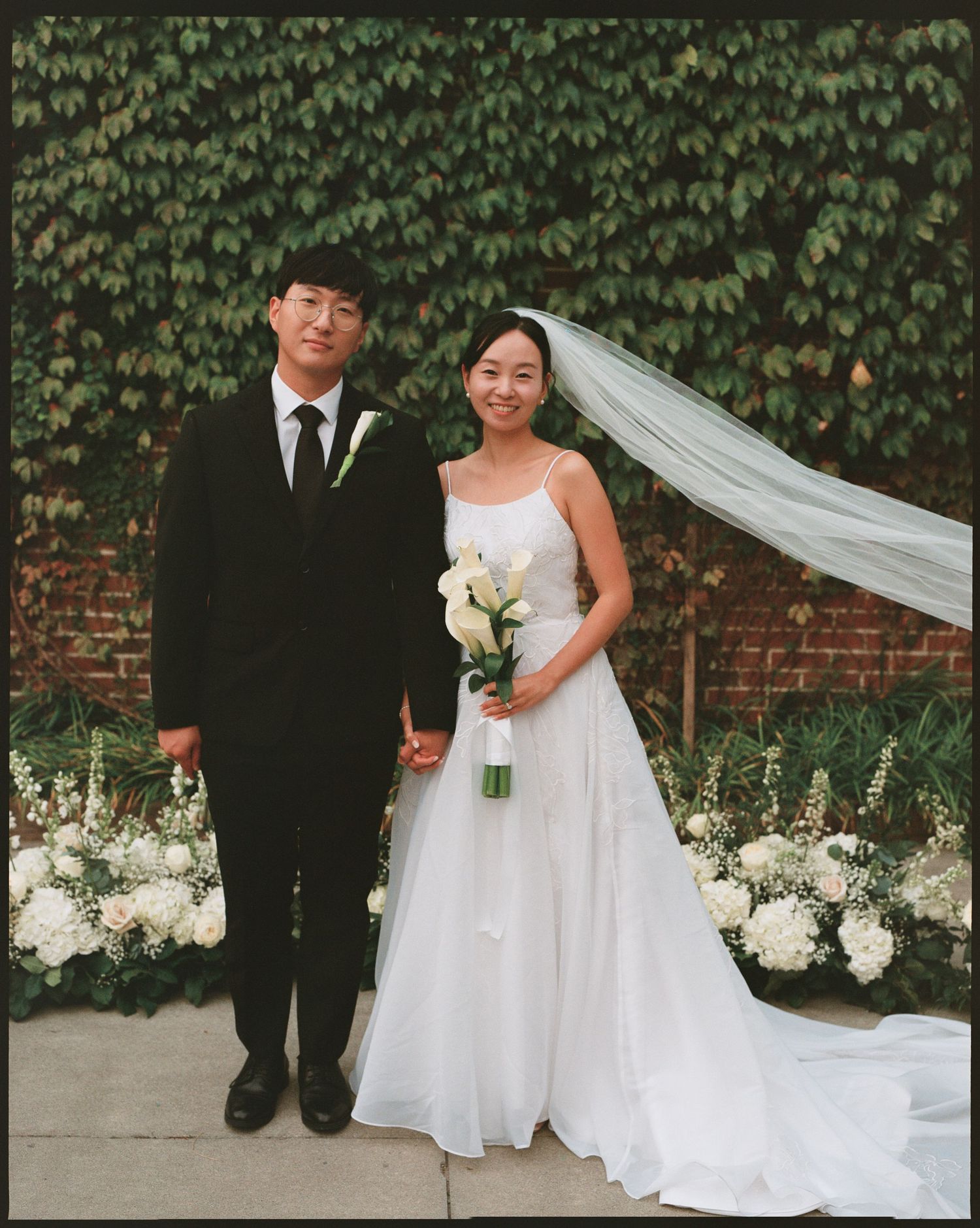 Wedding couple posing against a brick wall with lush green ivy backdrop.