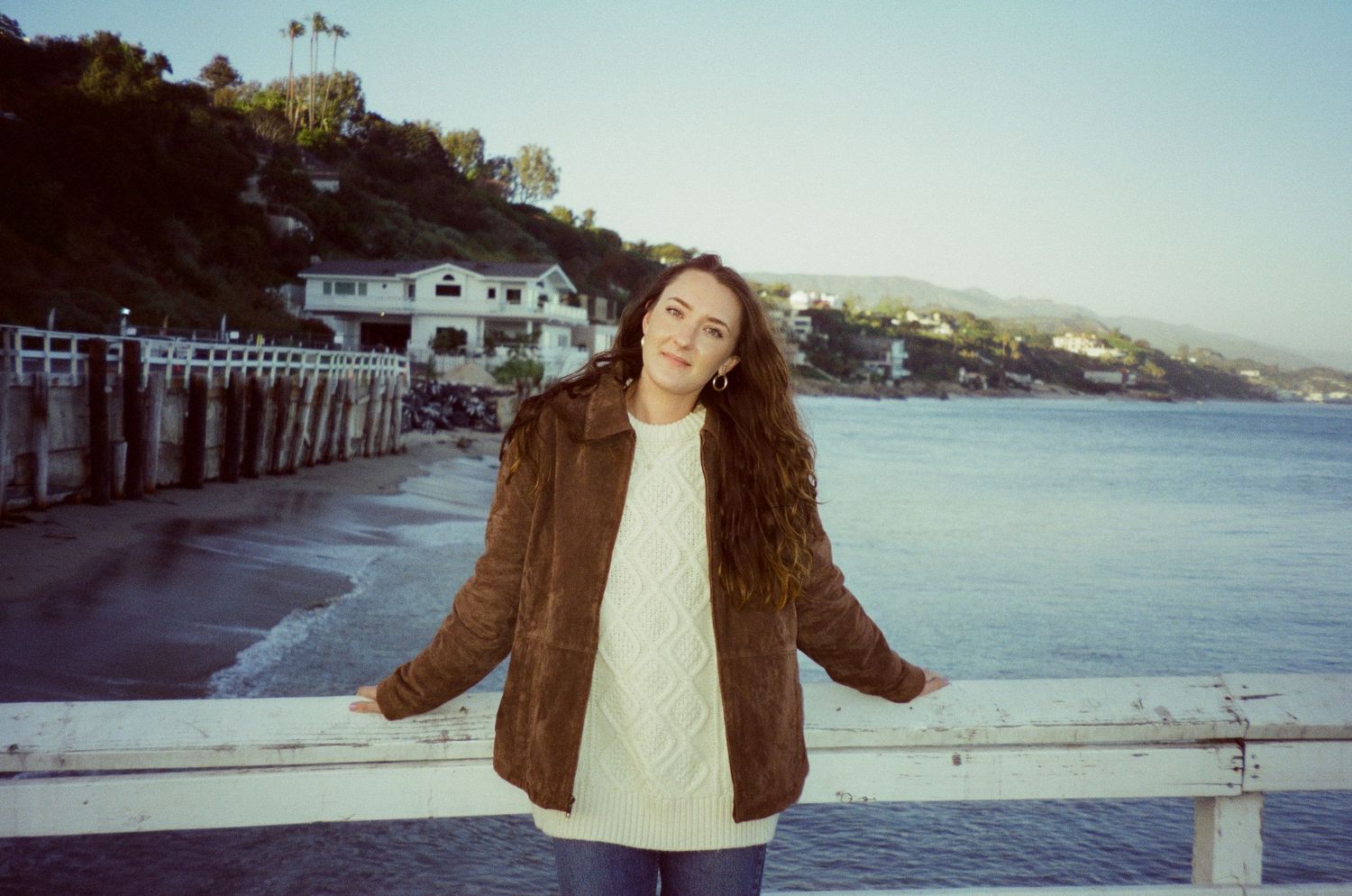 A scenic coastal view shows a person enjoying a sunny day by the waterfront with mountains in the background.
