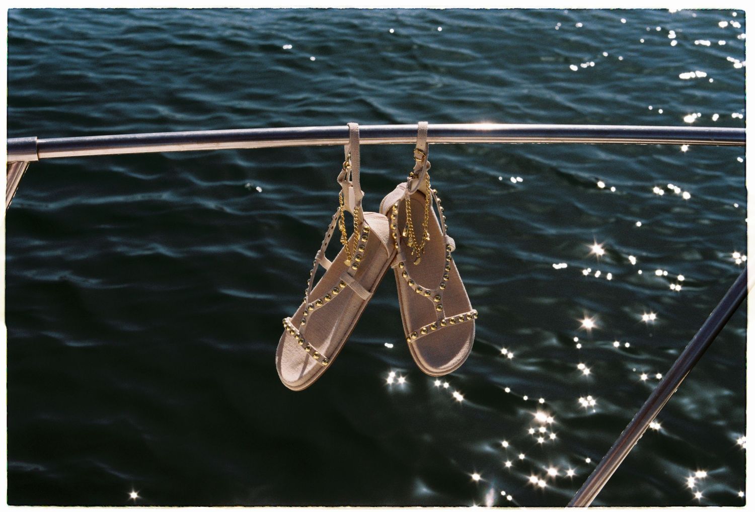 Gold studded sandals hanging on a boat railing over sparkling blue ocean water.