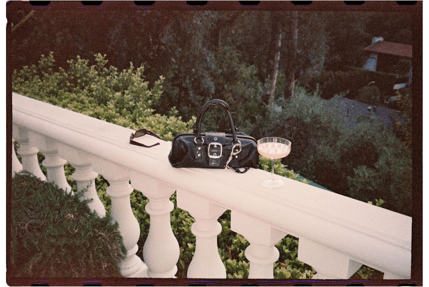 Black leather handbag and wine glass on white balcony railing with lush green foliage in background.