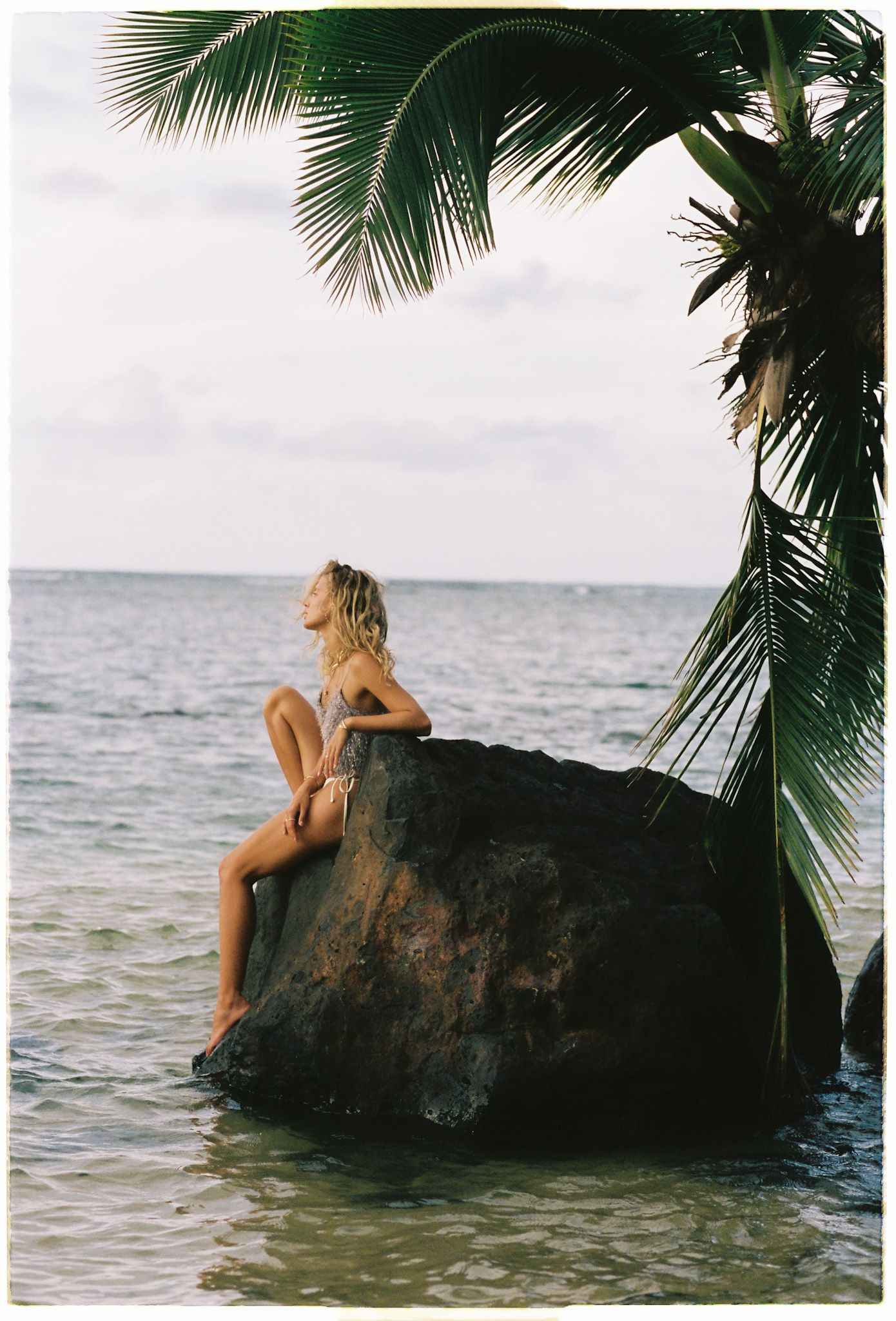Woman with blonde hair sits on a large dark rock in calm tropical water beneath a overhanging palm frond at sunset.