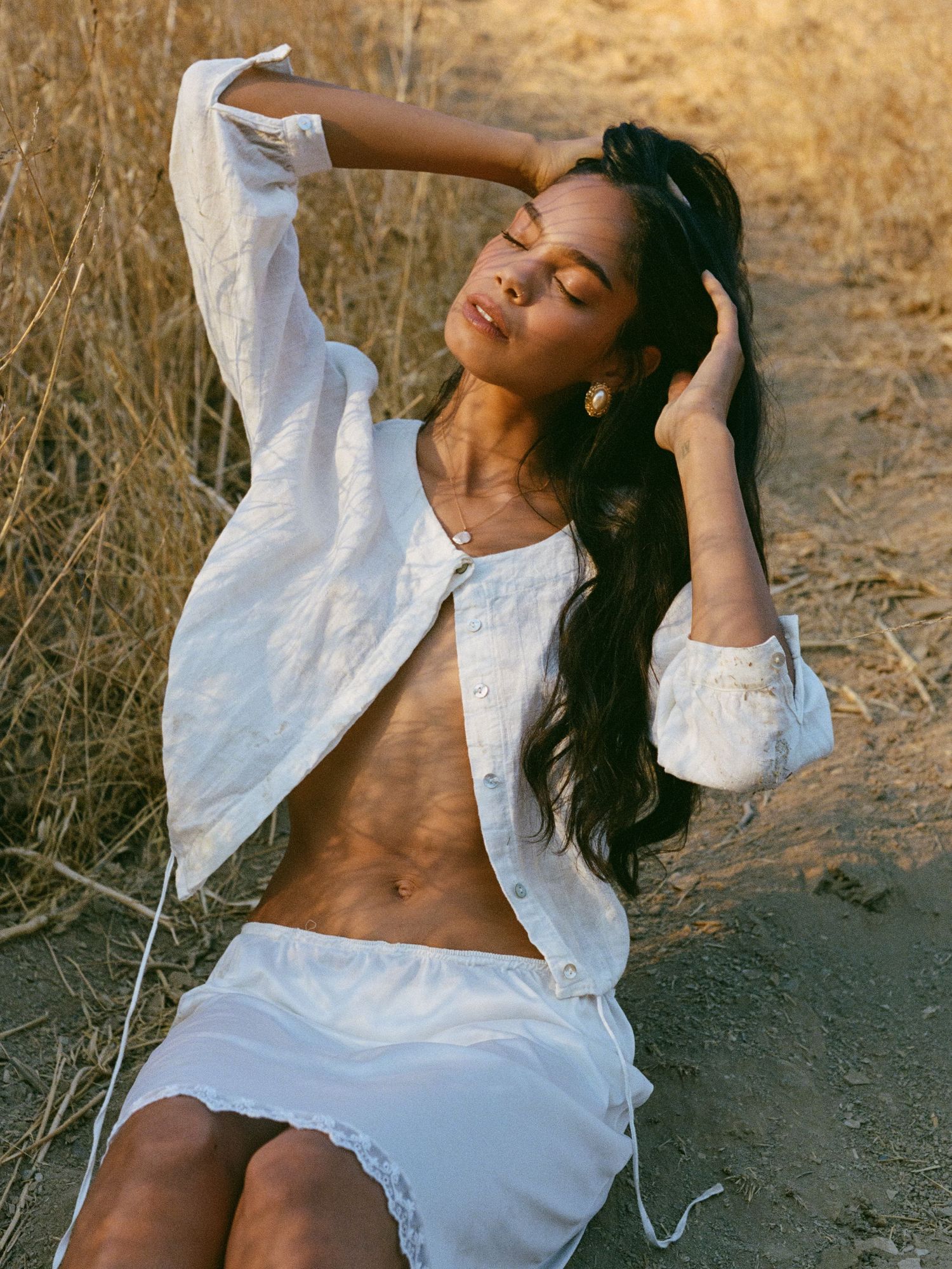 Model in white dress poses dramatically on ground during golden hour.