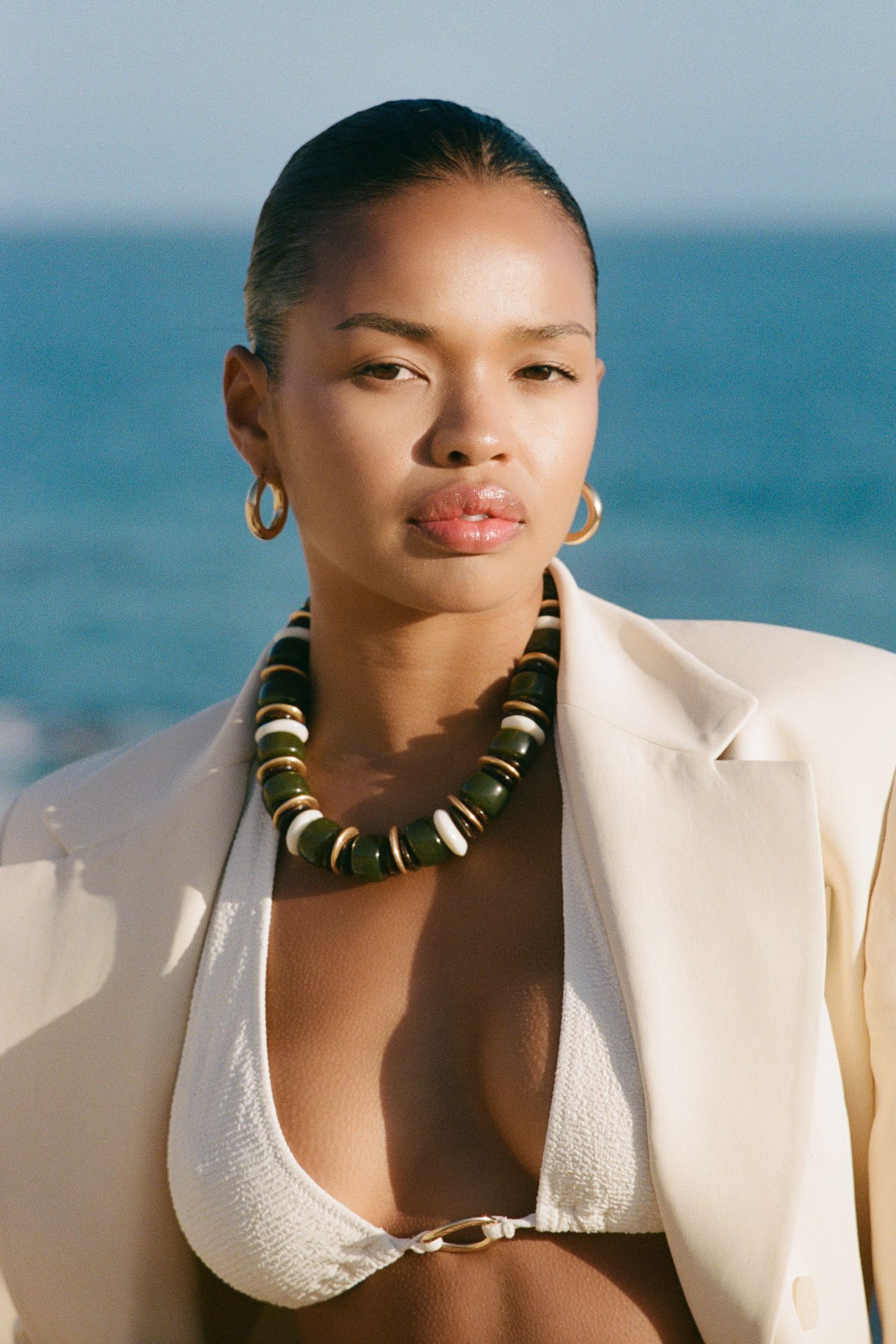 Fashion photo shoot at beach with model wearing white blazer and statement necklace.