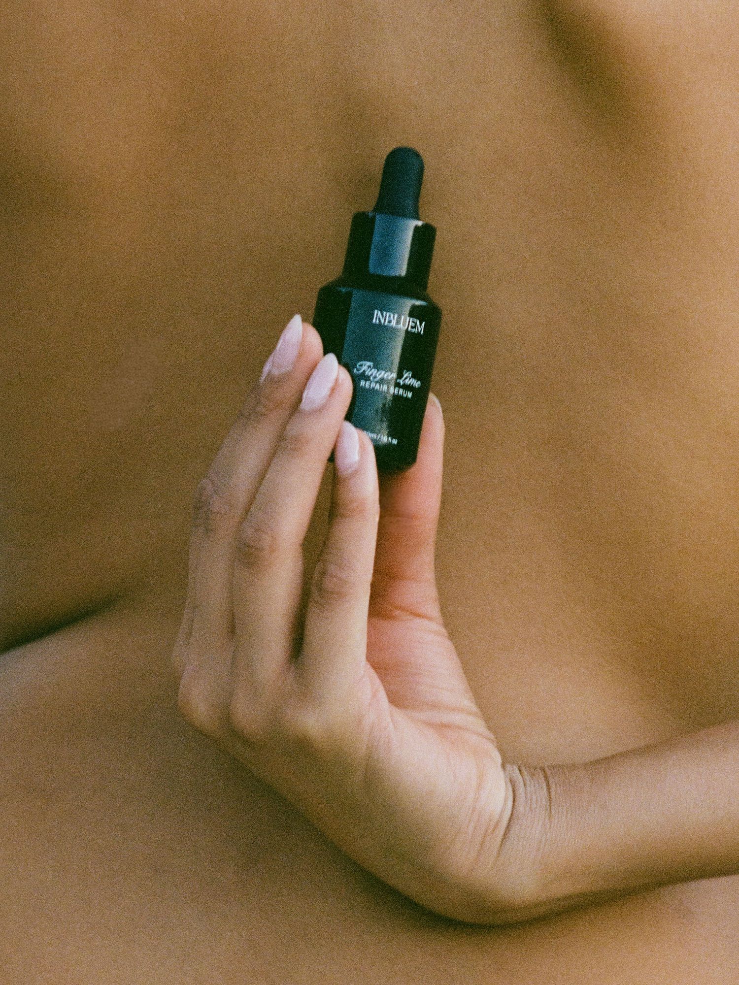 Hand holding a dark glass dropper bottle of skincare serum against a neutral background.