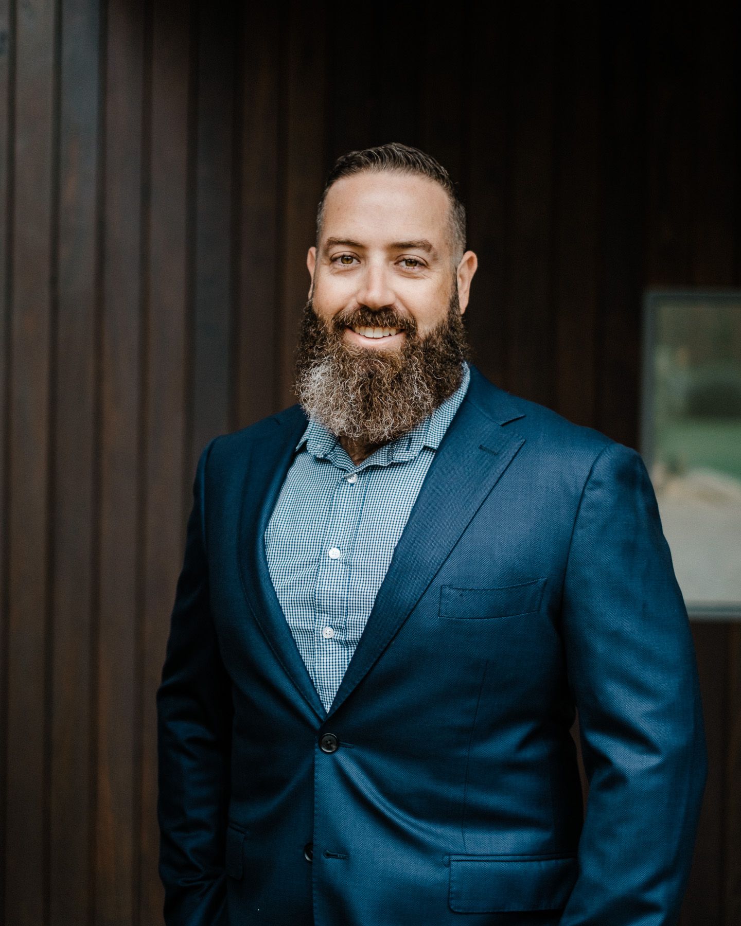 A bearded man in a navy suit and light blue shirt poses against a dark wooden wall in a professional headshot series.