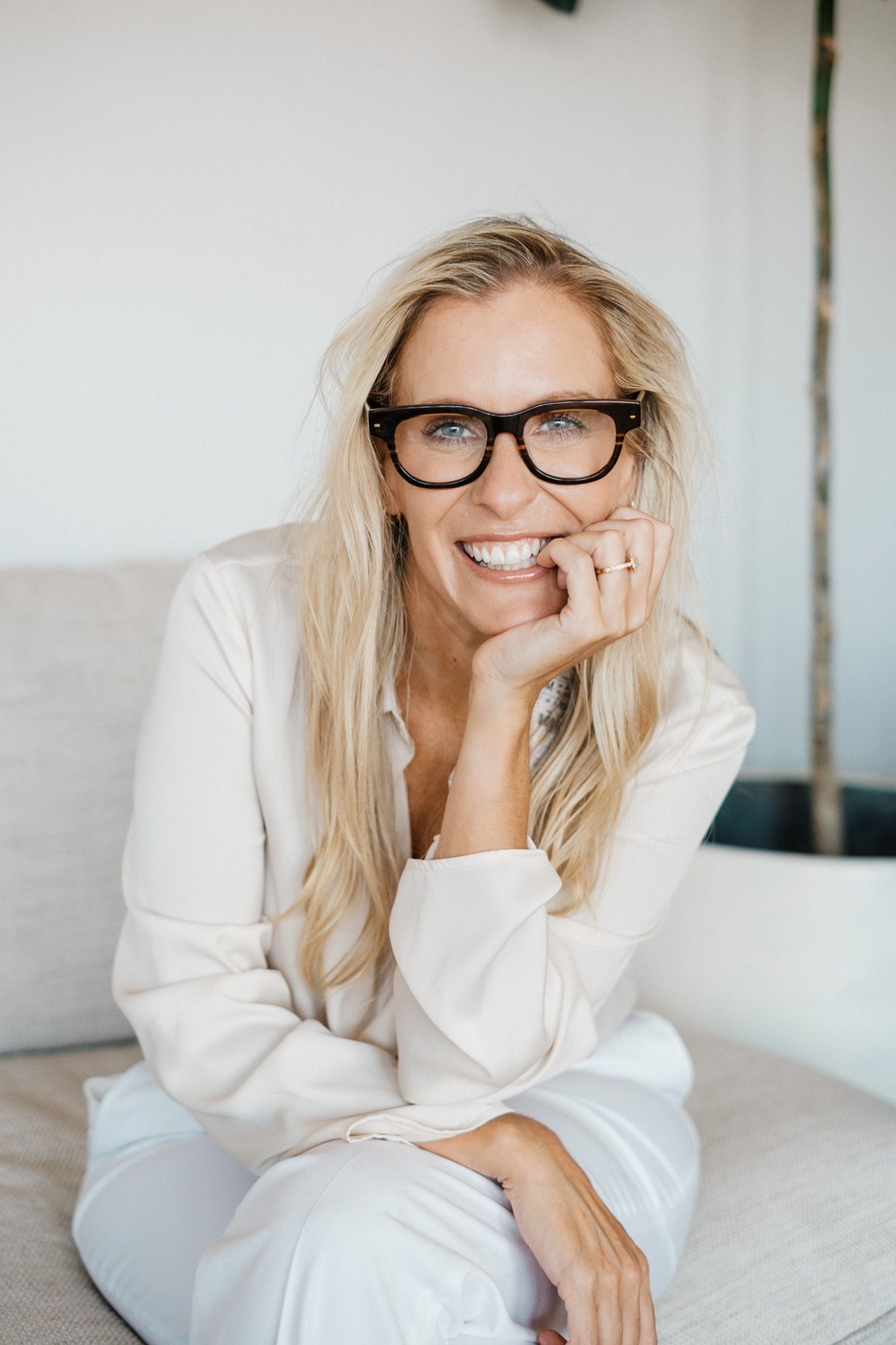 Professional lifestyle portrait of someone with blonde hair and glasses relaxing in casual white attire on white bedding.