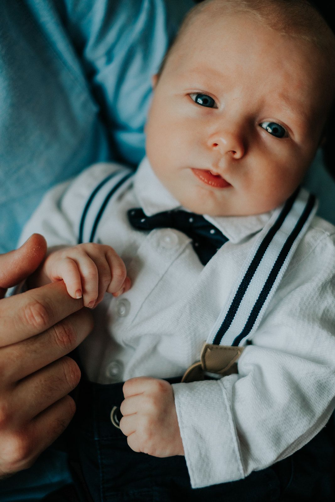 A baby wearing a white dress shirt with black suspenders and bow tie lies on turquoise fabric while holding someone's finger.