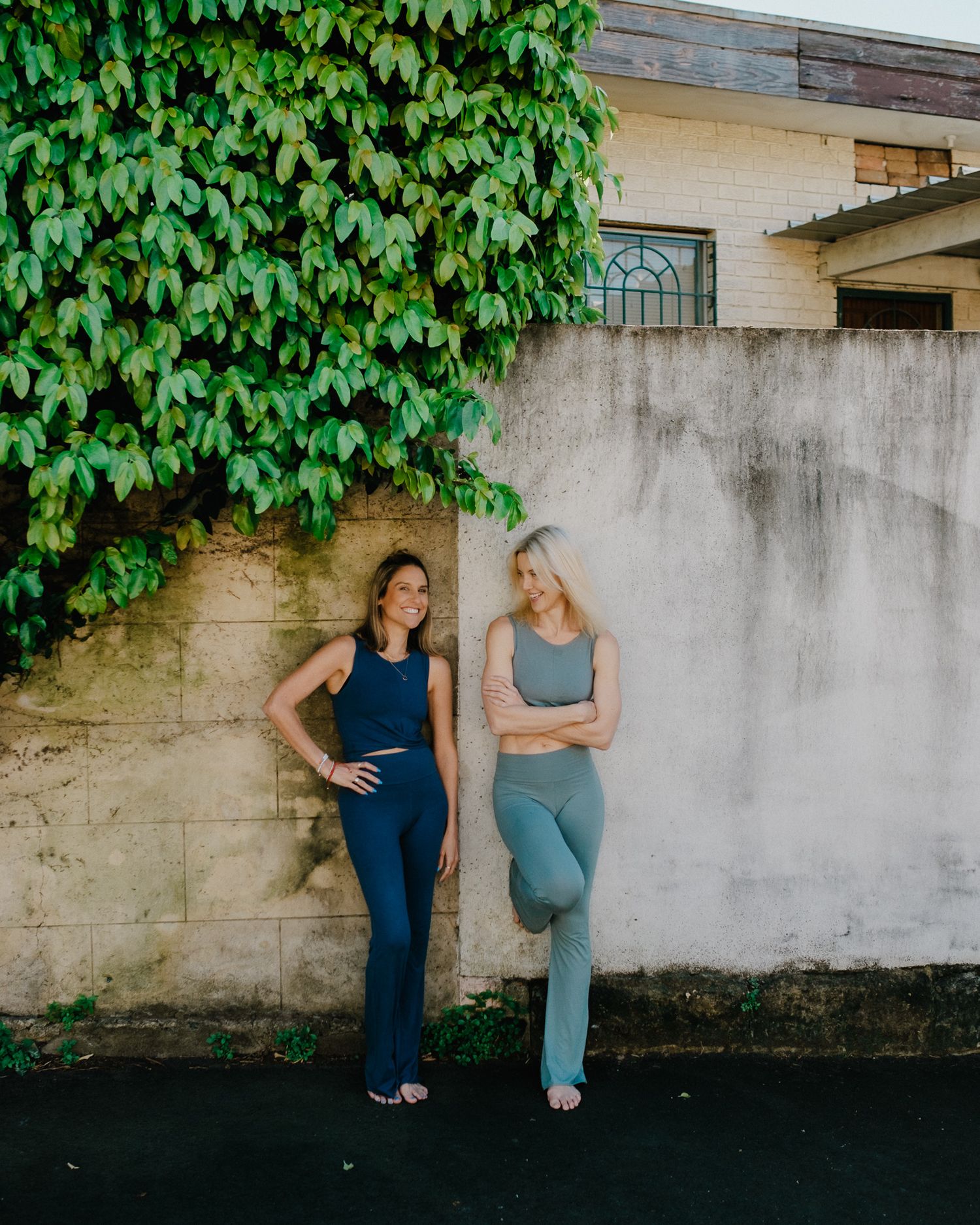 Two women in workout attire pose together against a concrete wall with green ivy growing above.
