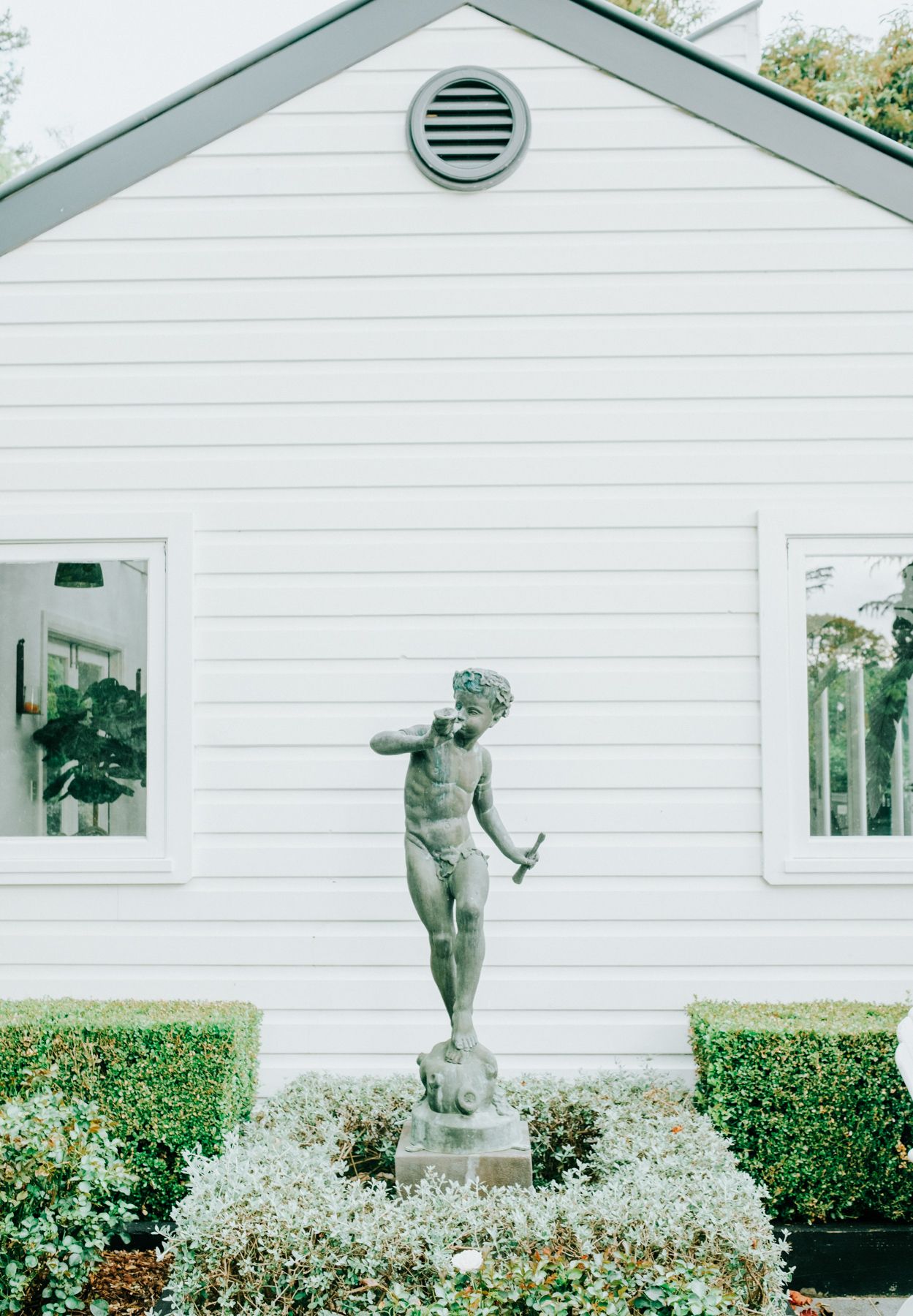 A classical nude statue stands in a garden bed against a white house with symmetrical windows and a circular vent.