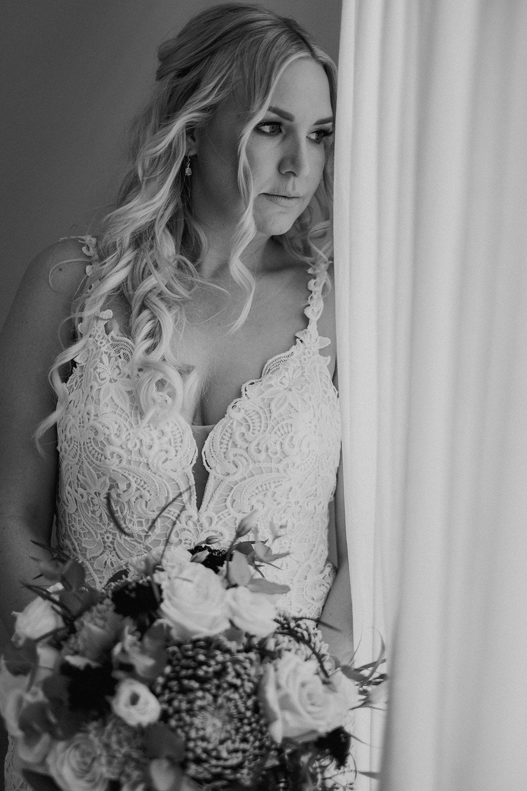 A bride in a white lace wedding dress holds a bouquet of roses and baby's breath against a light backdrop.