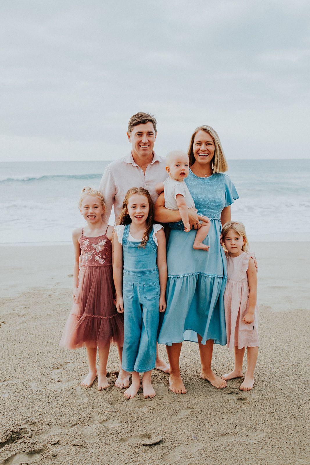 Family wearing coordinated turquoise and pink outfits poses together on a sandy beach at sunset.
