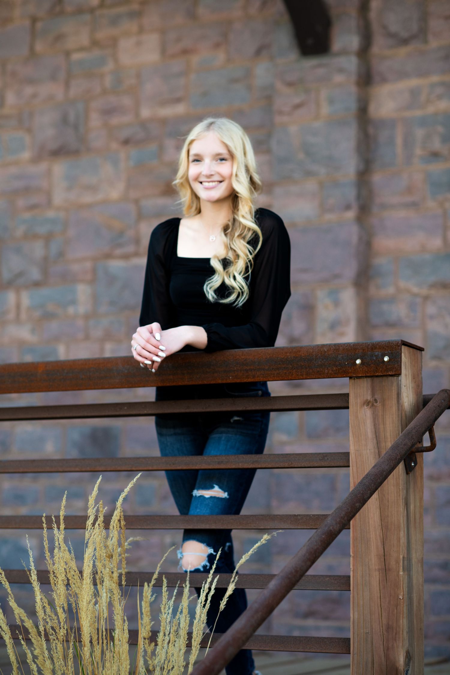 Woman in black sweater poses by wooden railing with stone wall backdrop.