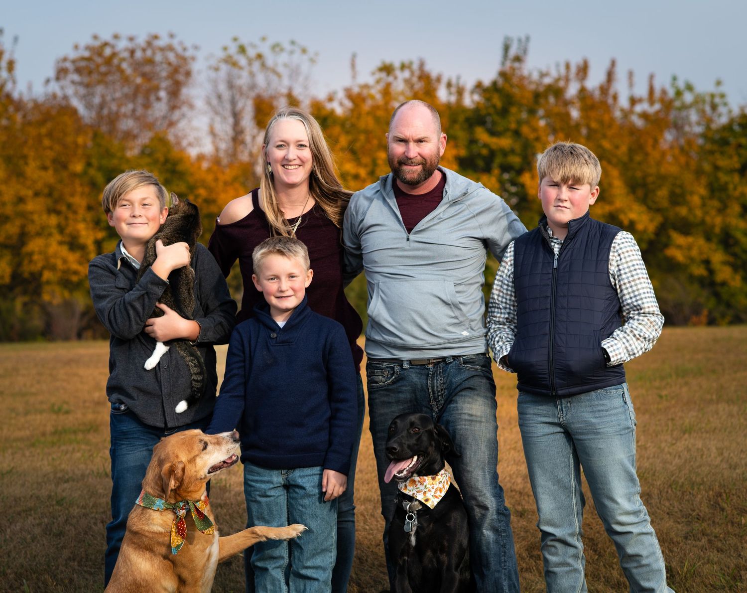 Family portrait featuring couple, three kids and two dogs standing together in a fall field.