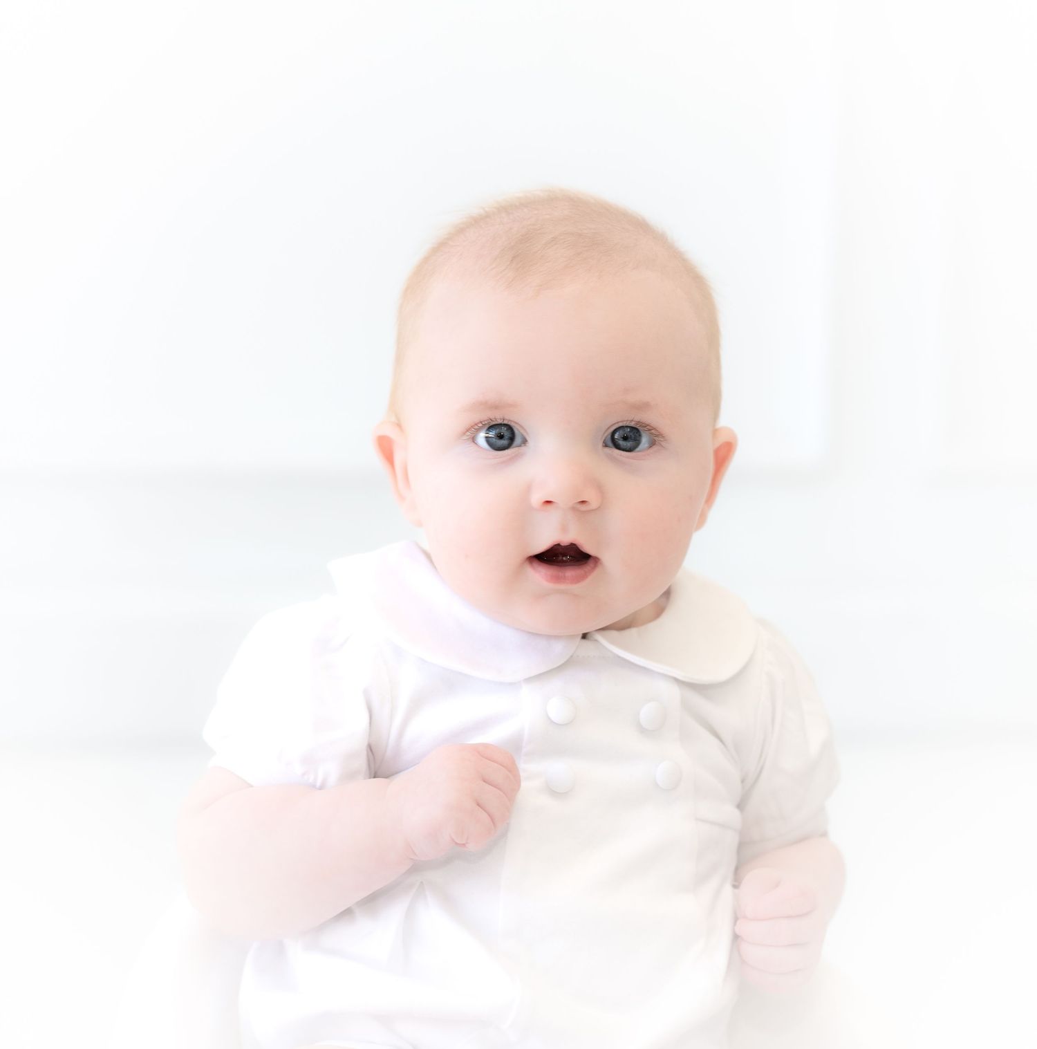 Baby in white outfit gazes upward with bright eyes against light background.