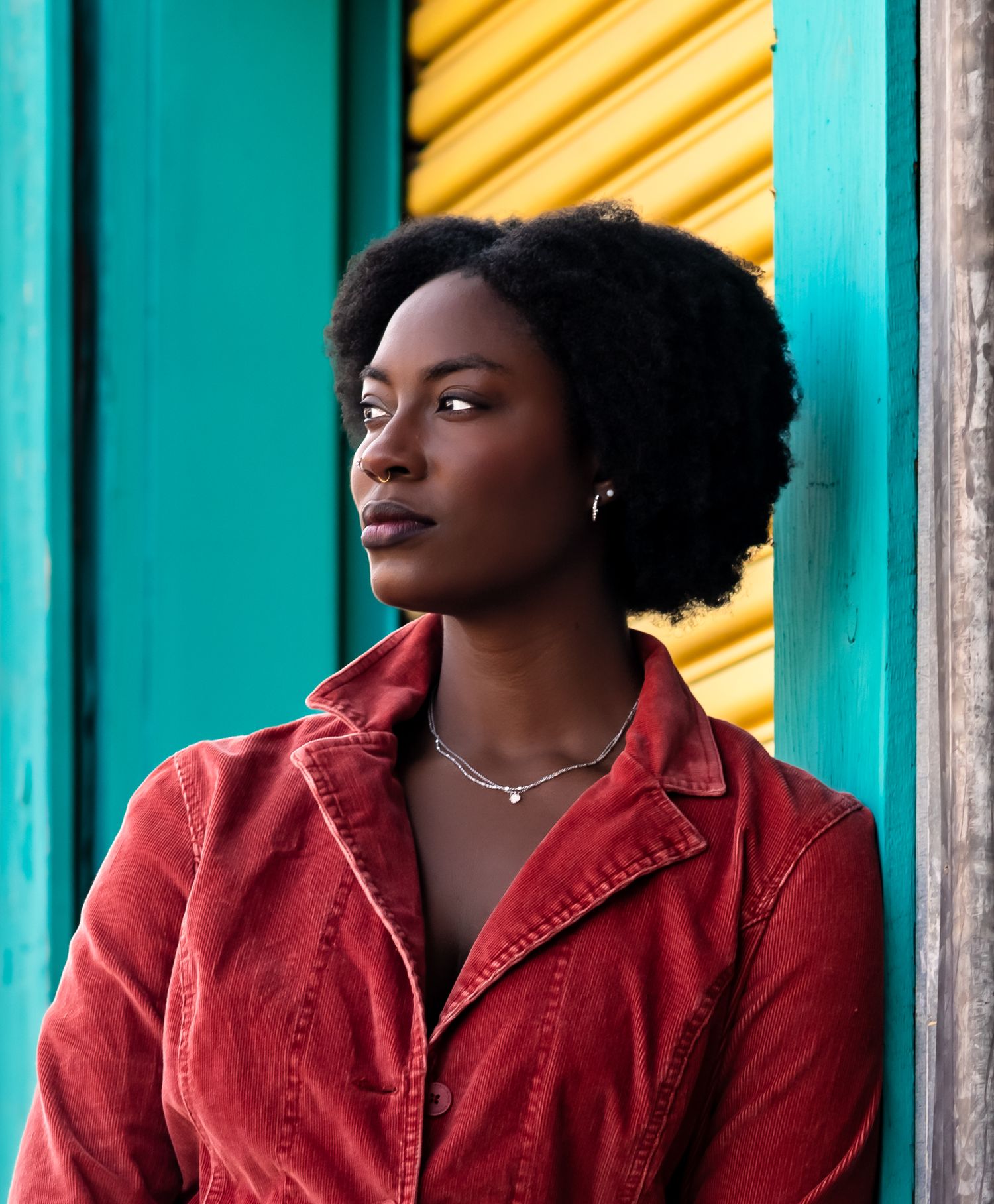 Portrait against turquoise wall and yellow shutters wearing red corduroy jacket and silver necklace.