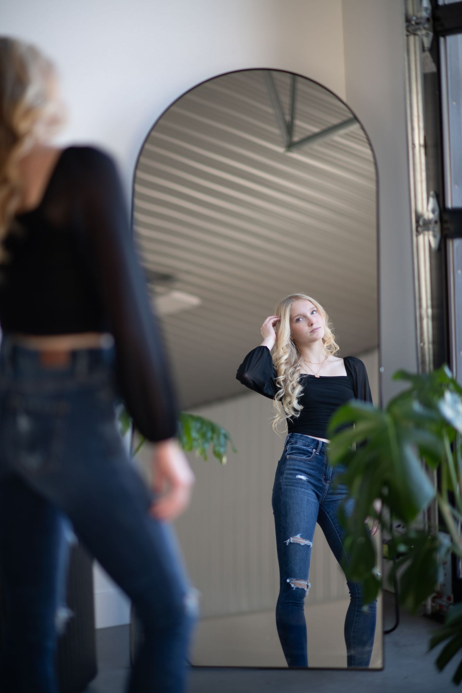 Mirror reflection shows person in black top and distressed jeans posing near indoor plants.