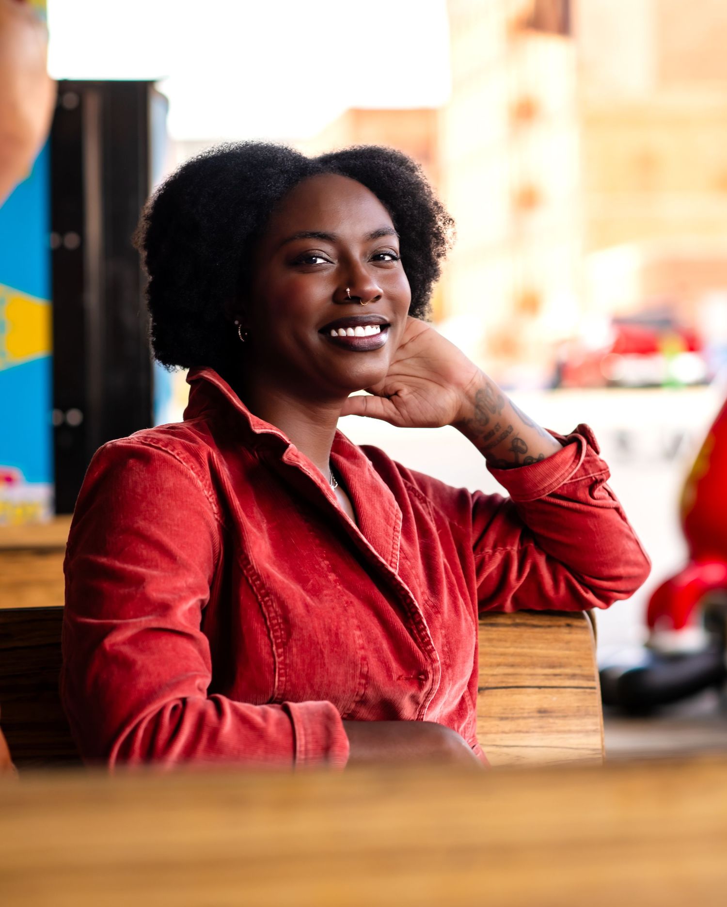 Person in red corduroy shirt sitting at wooden table with relaxed pose and natural curly hairstyle.