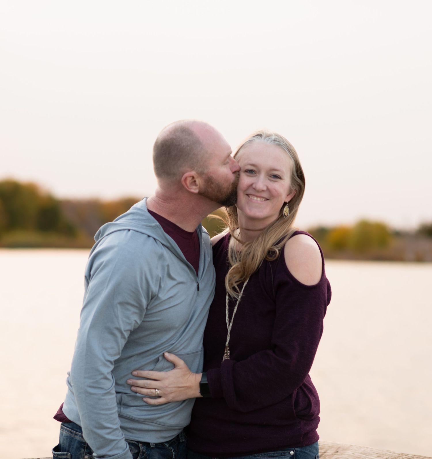 Loving couple shares an intimate moment by a lake during sunset with autumn foliage in the background.