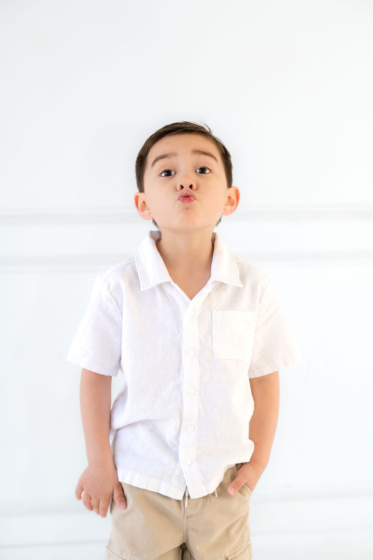 Young boy in white shirt and khaki shorts making playful face against white background.