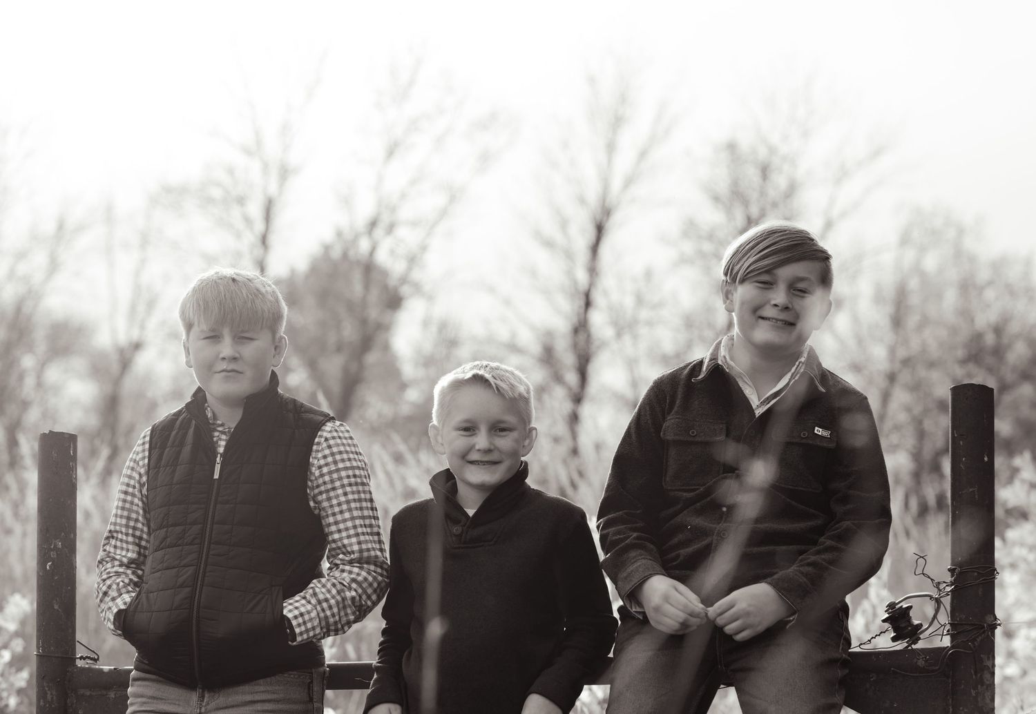 Three siblings sit together on a wooden fence in a black and white vintage-style photograph.