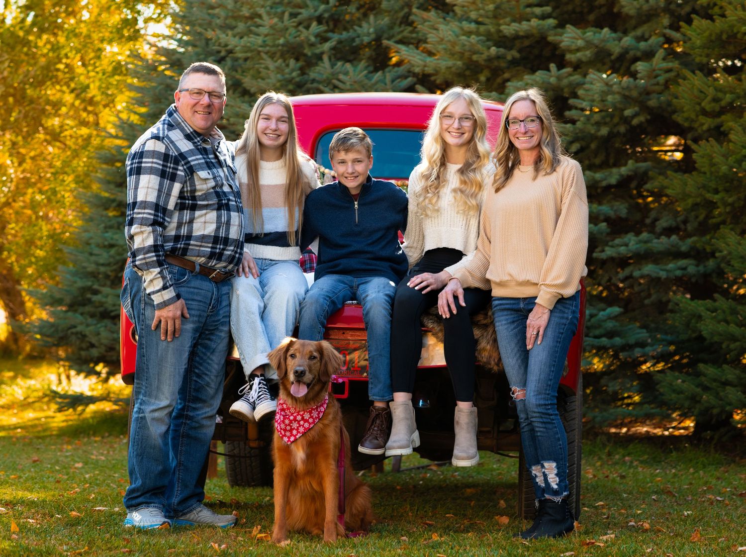 Family poses with their brown dog in front of a classic red pickup truck surrounded by autumn trees.