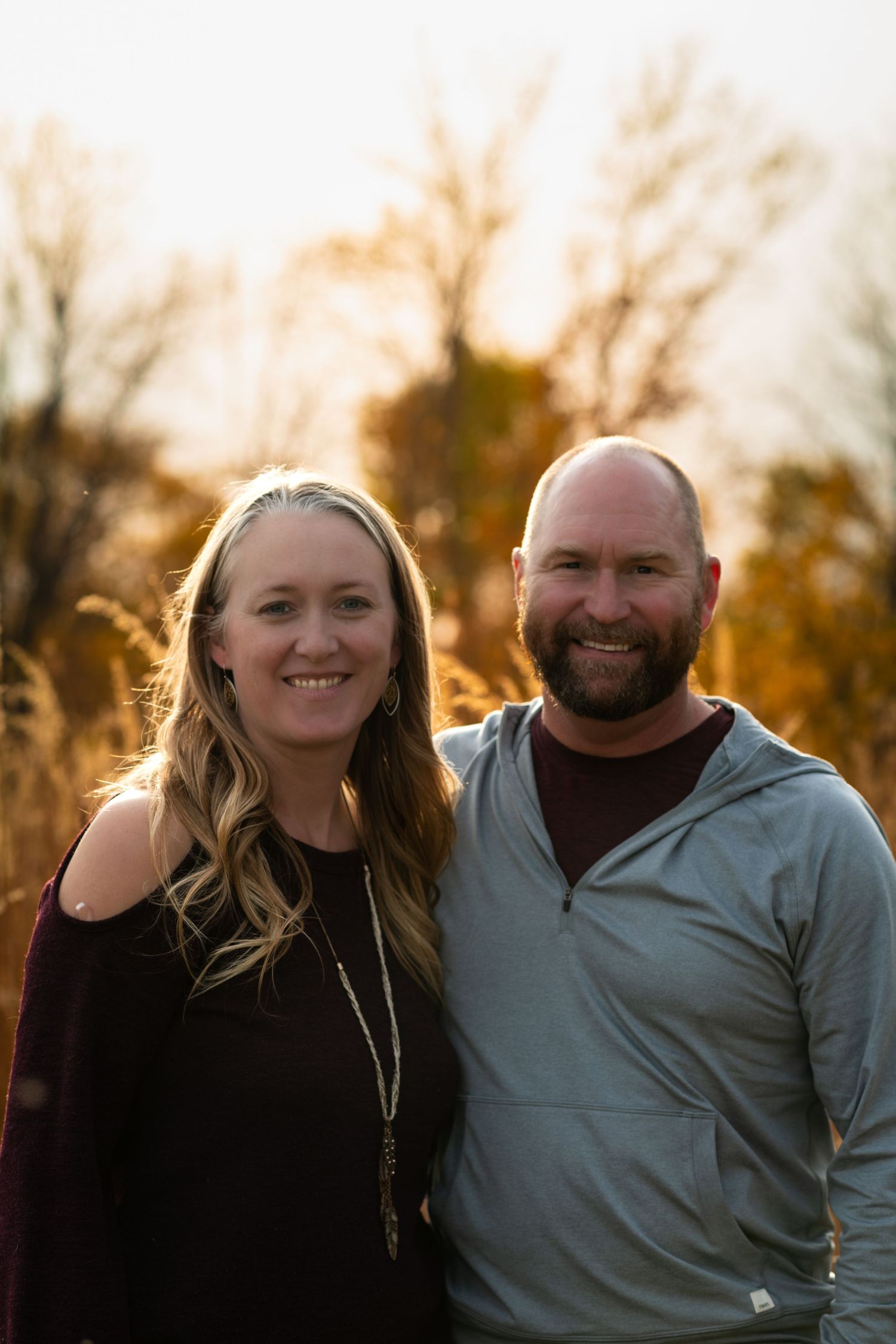 Couple poses together outdoors during golden hour with fall trees in background.