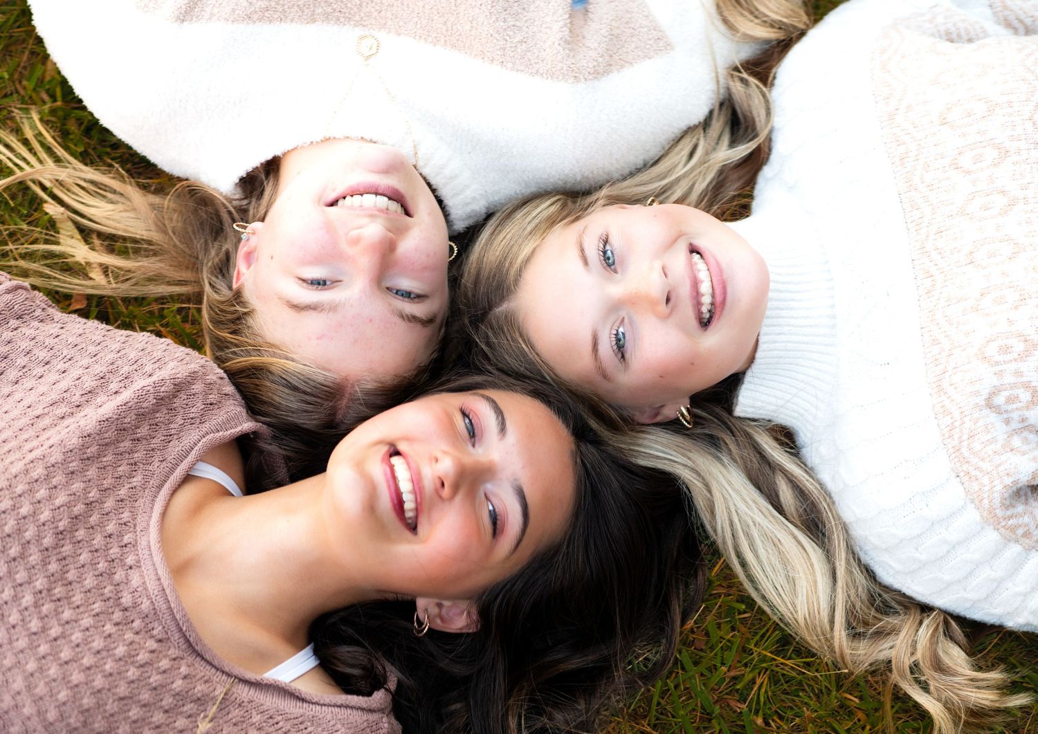 Three friends lying on grass wearing sweaters share laughter together in natural light.