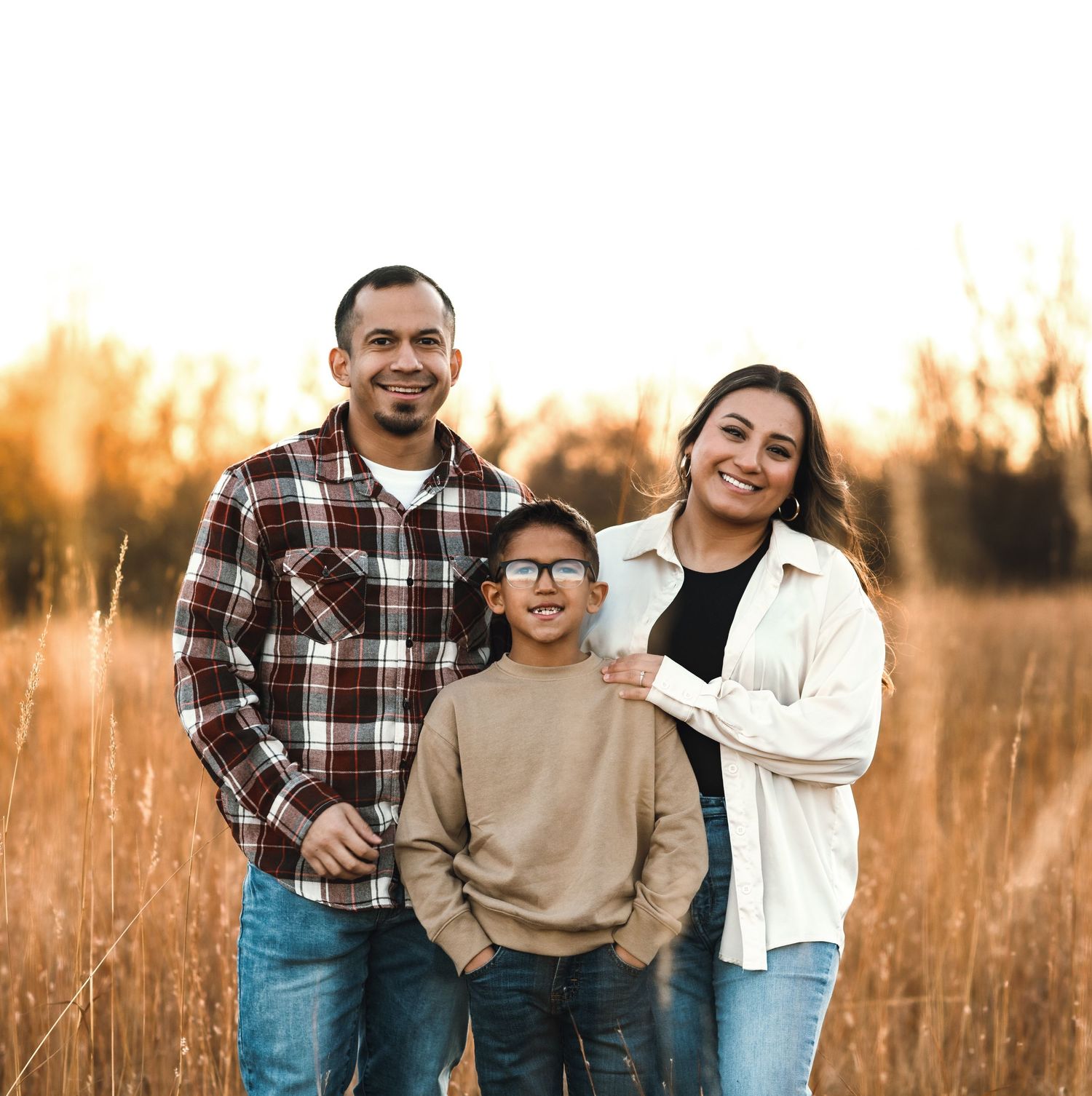 Family of three poses together in a golden field at sunset wearing casual attire.