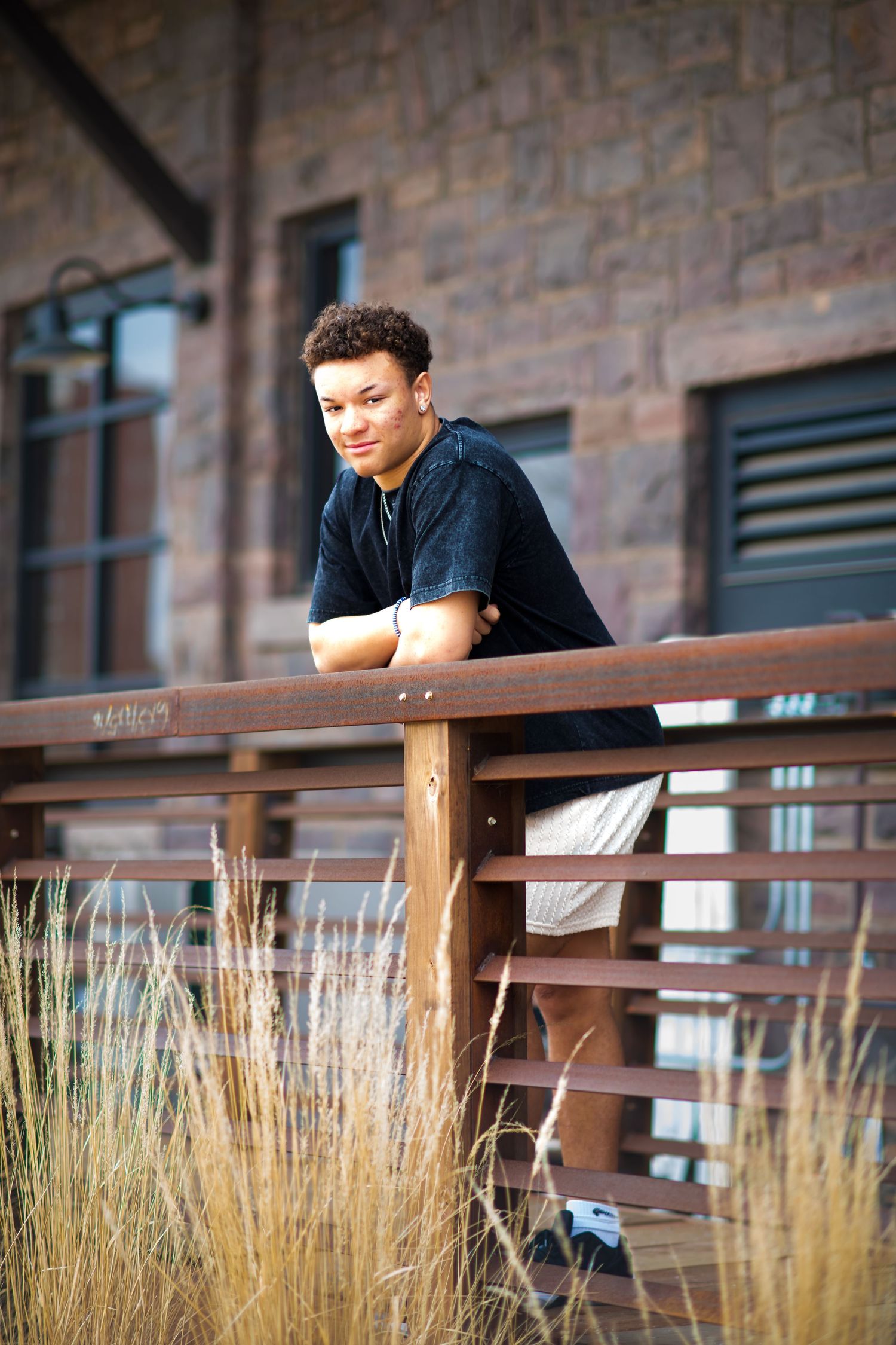 Young person in black shirt leans on wooden railing with ornamental grasses in foreground during outdoor portrait session.