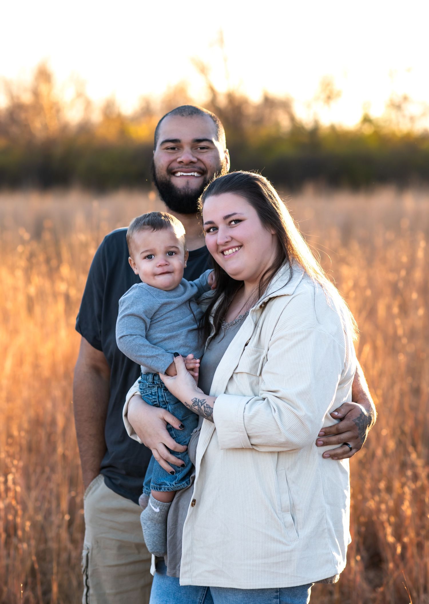 Family of three poses together in field during golden hour sunset.