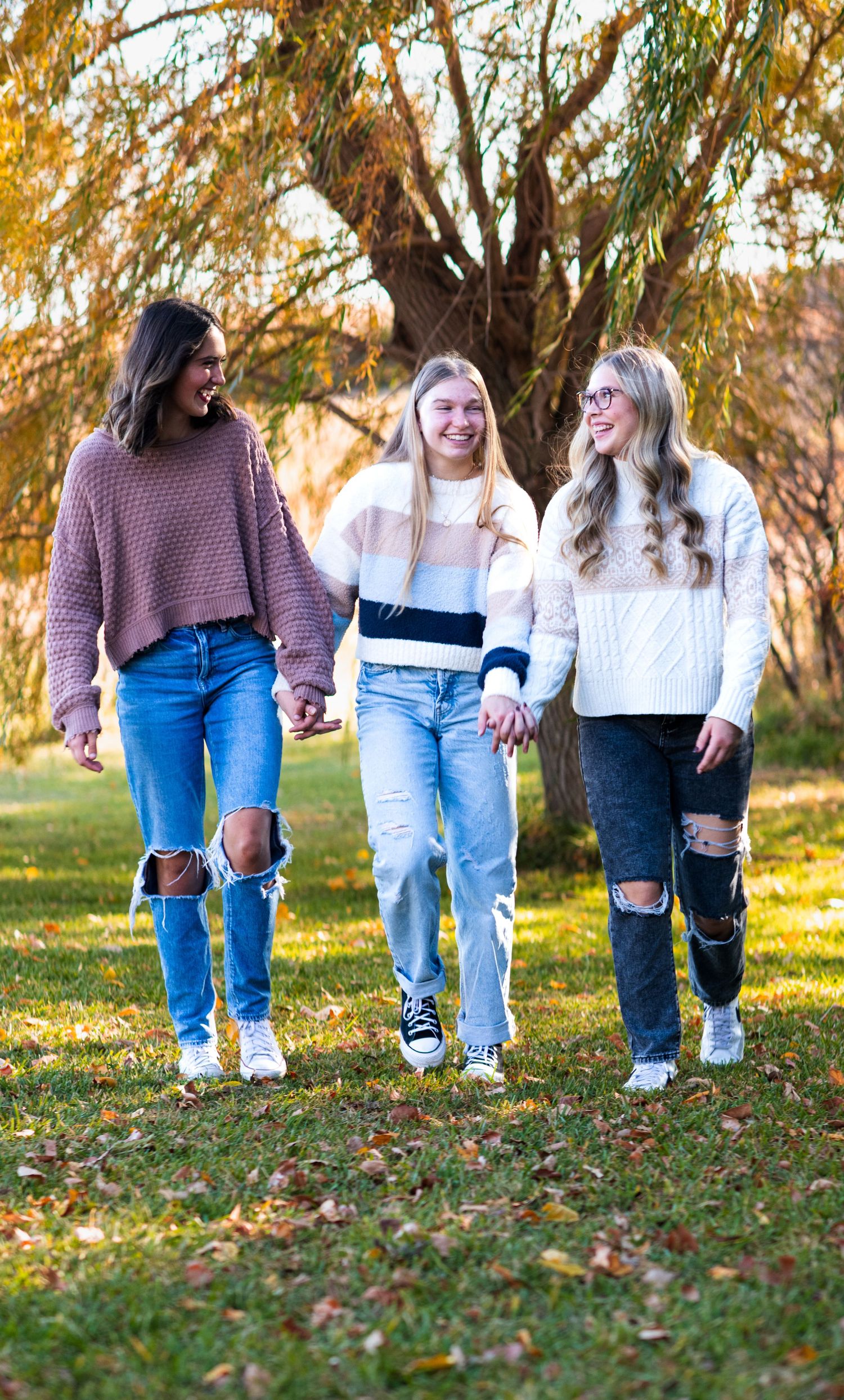 Three friends in cozy sweaters walk together through fallen autumn leaves.