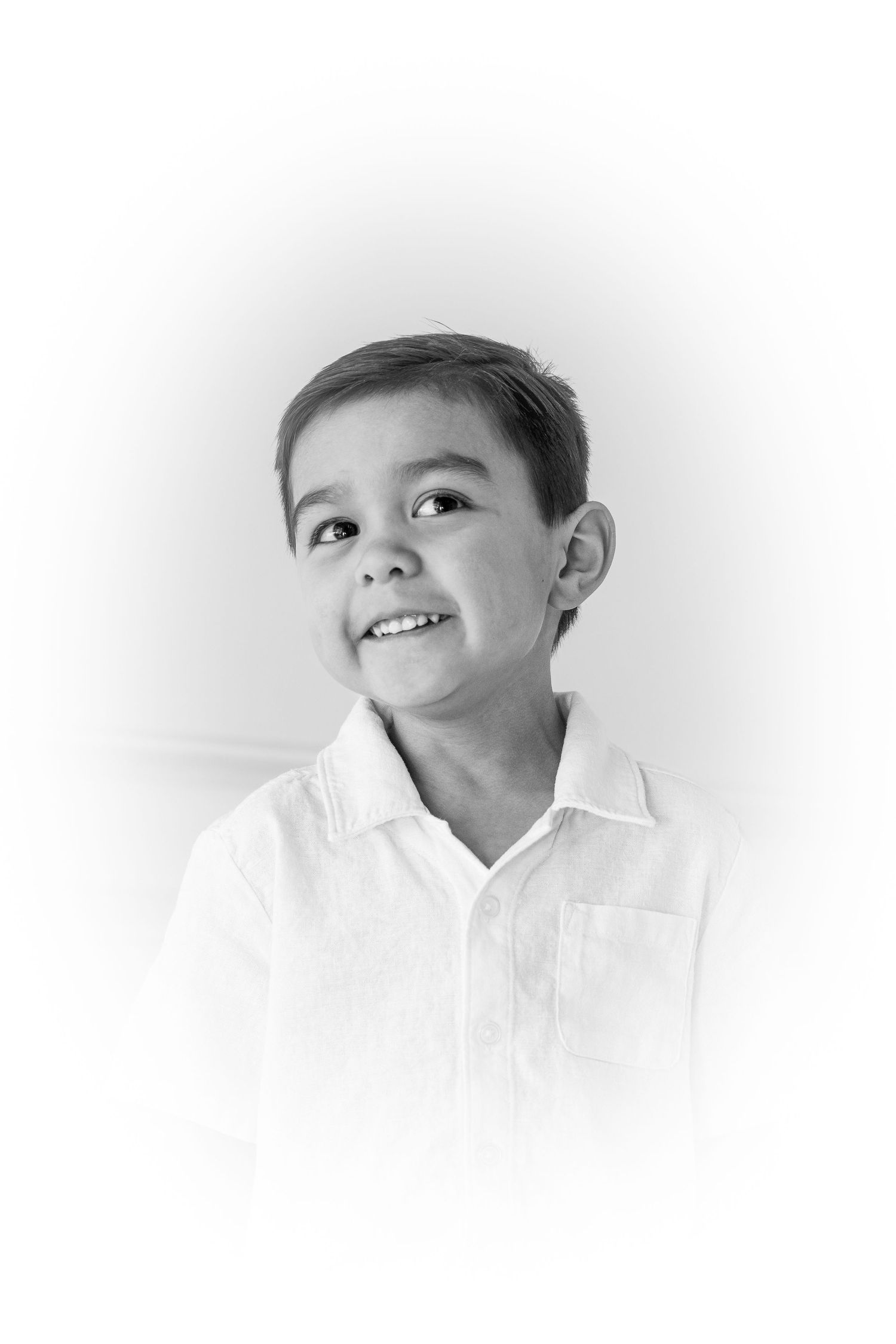 Black and white studio portrait of young child in white collared shirt smiling upward.