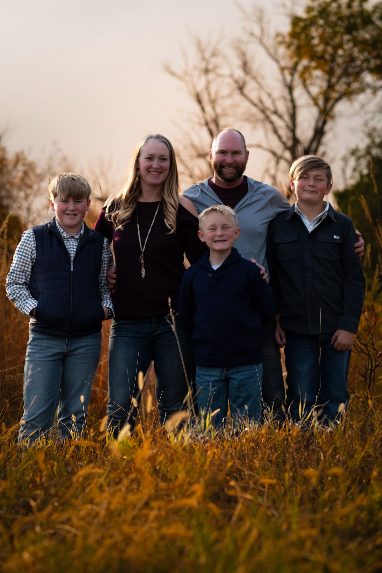 Family of five poses together in tall grass during sunset with autumn colors in background.
