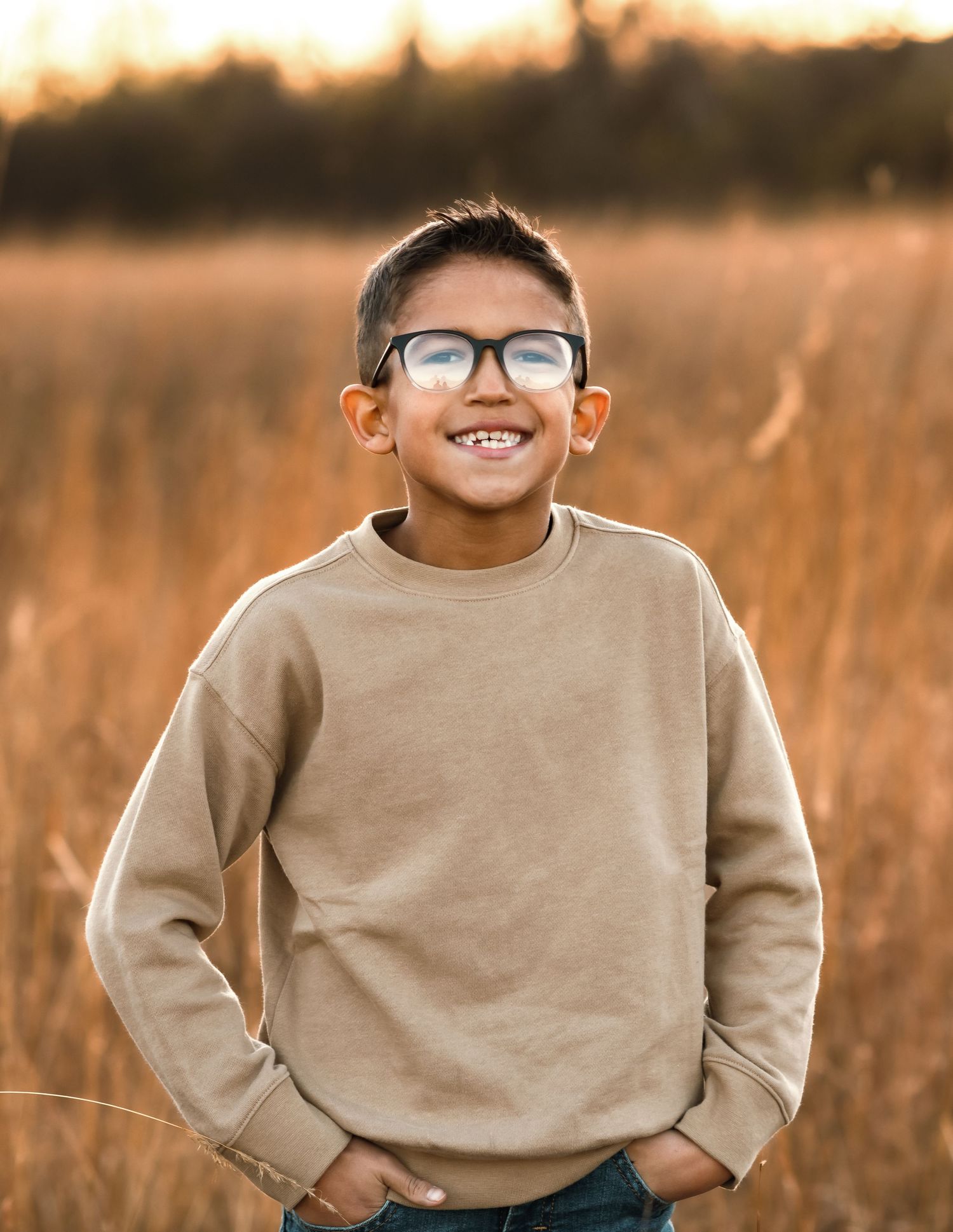 Young person in beige sweater smiles brightly wearing glasses in field at sunset.