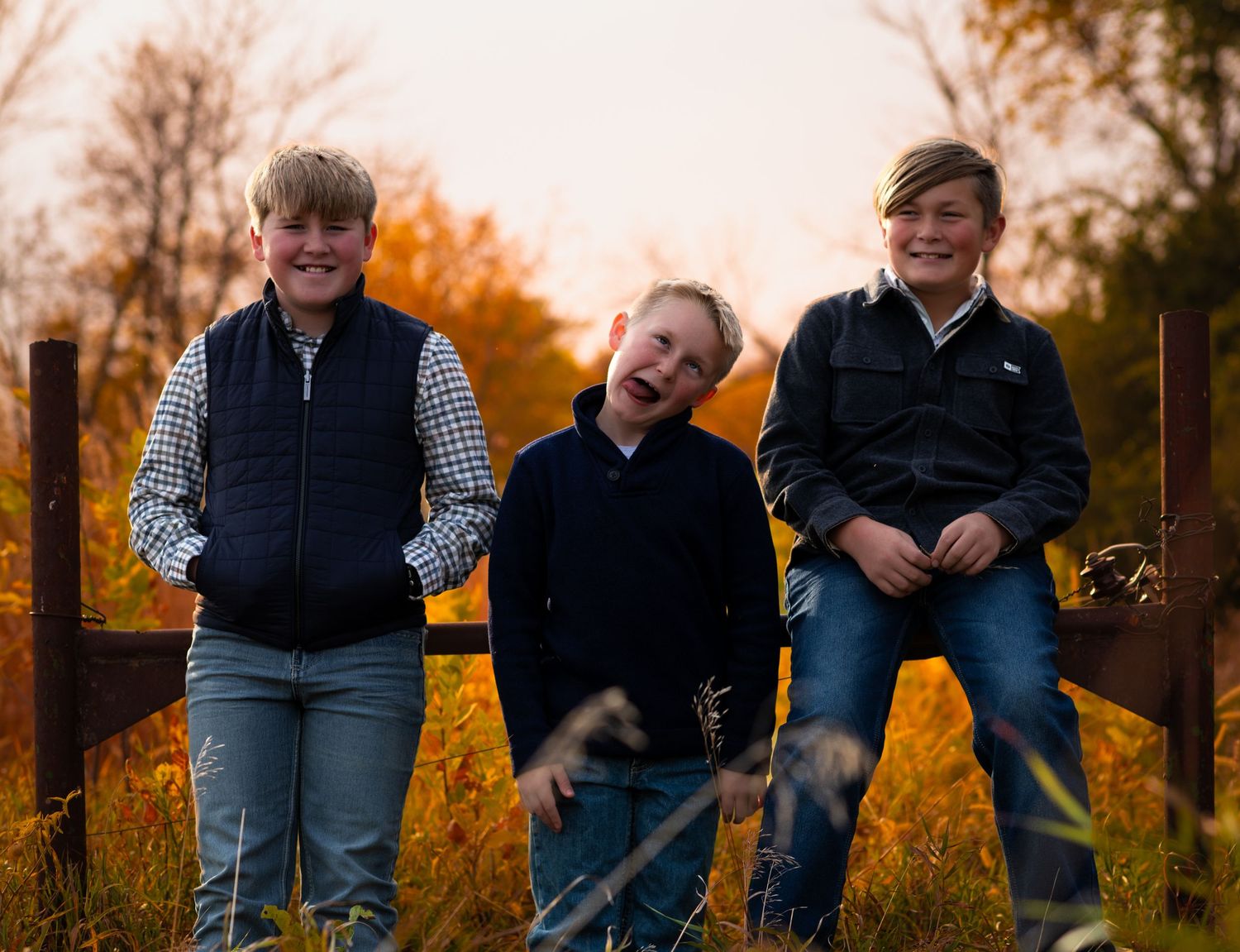 Three children sitting on wooden fence railing while laughing together in autumn setting.