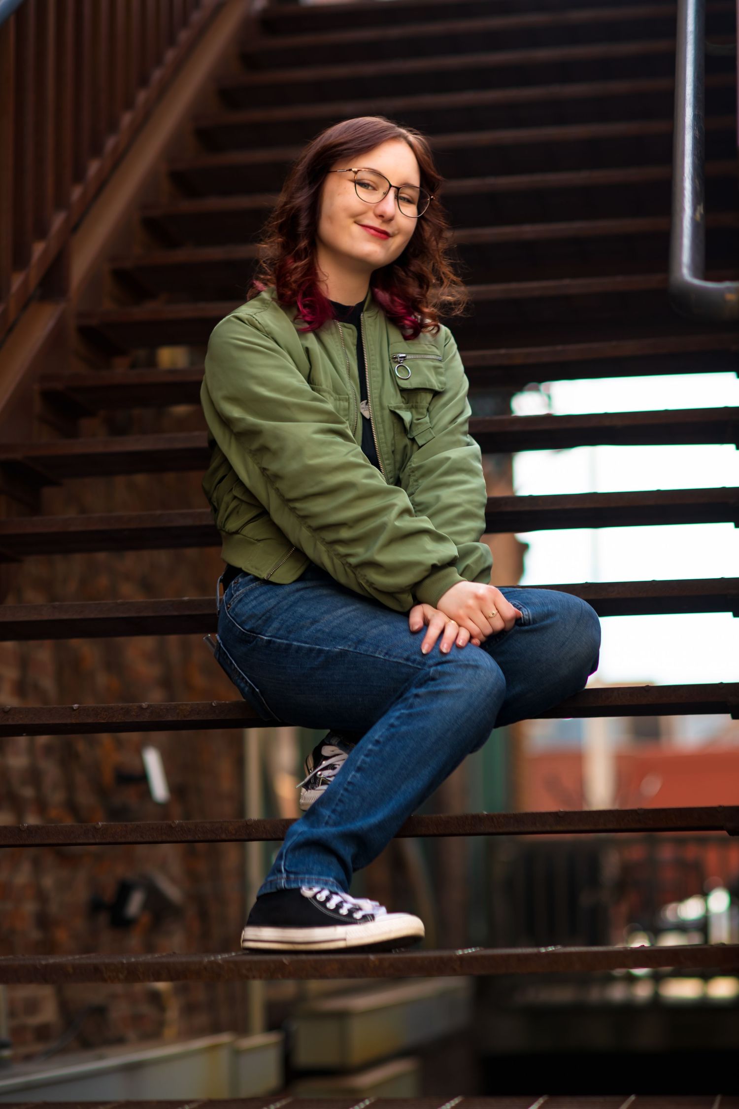 Person in green jacket and jeans sits casually on industrial metal stairs.