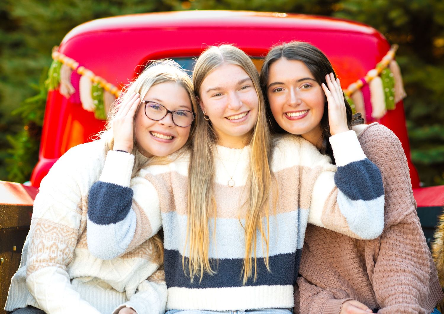 Three friends in cozy sweaters sit together and smile joyfully against a red backdrop outdoors.